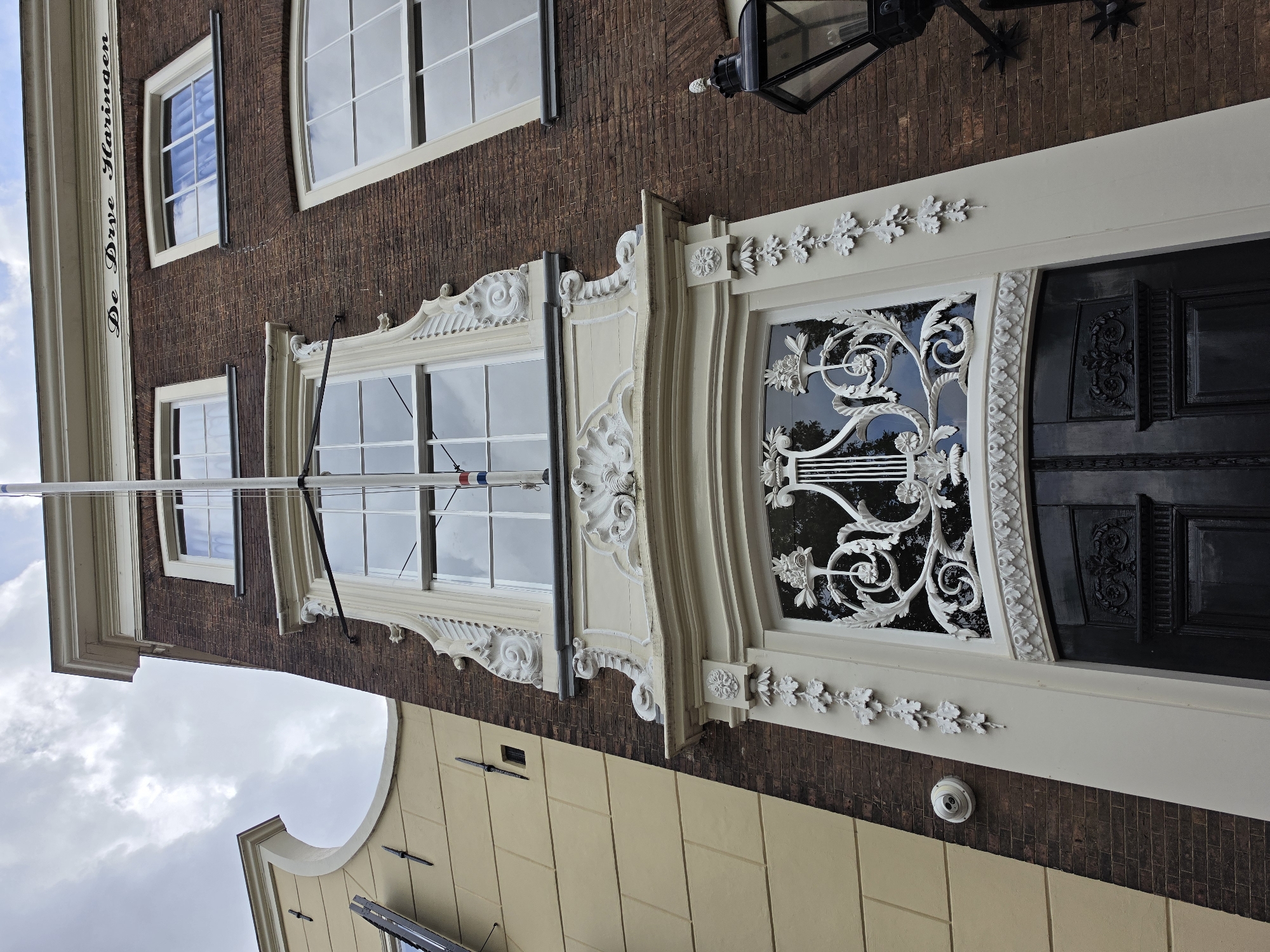 The image shows the facade of a brick building with ornate white decorative elements above a dark wooden door. The building is located in Delft, Netherlands. The facade features a large arched window with intricate white detailing above a smaller, decorative grille that covers a window above the door. The door itself is dark and appears to be made of wood with carved panels. A flag pole extends vertically along the front of the building, with a small Dutch flag visible near the top. The building to the left has a pale yellow facade with a curved upper edge. The sky above is cloudy.