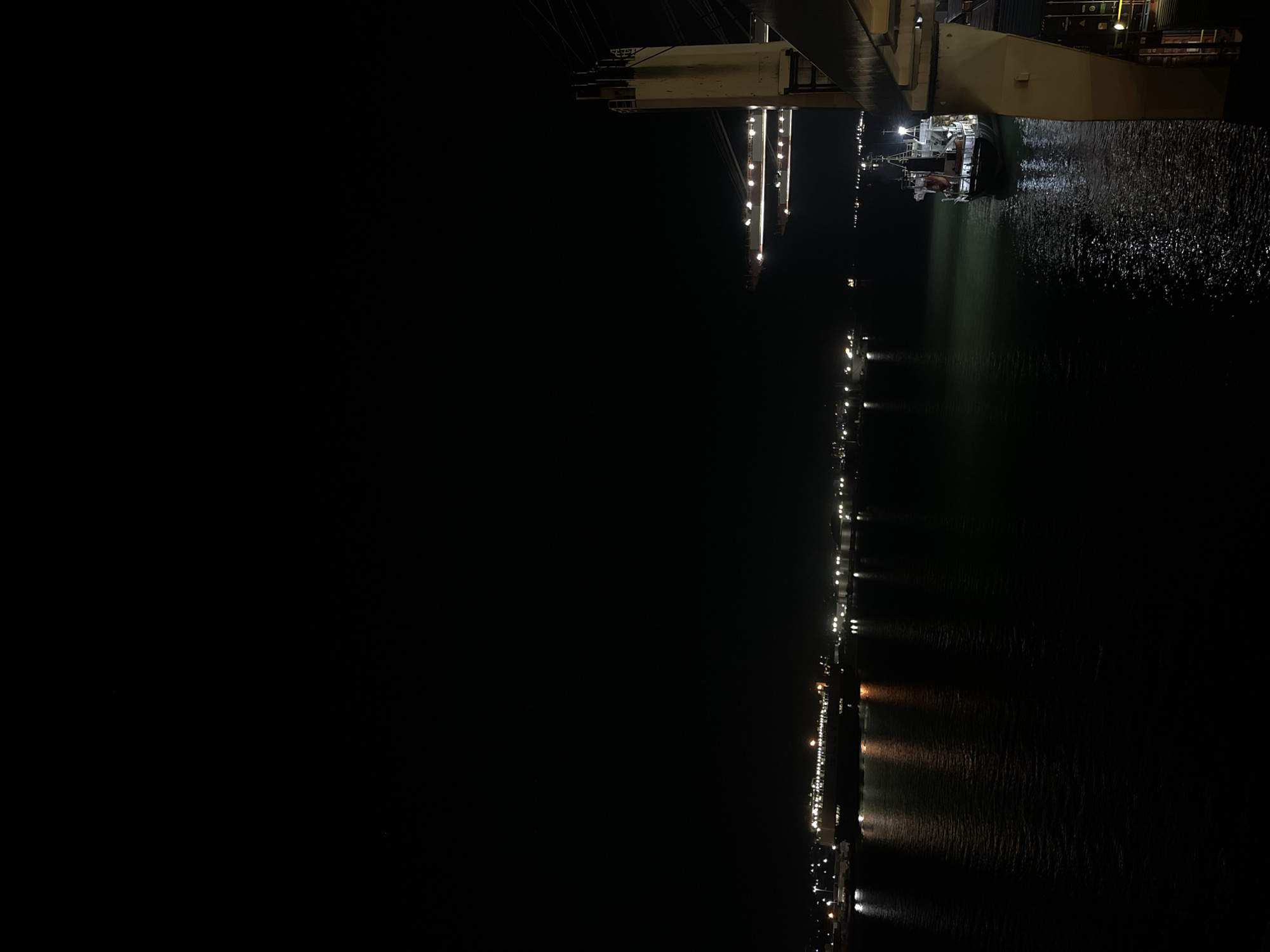 The image shows a night scene at a port or harbor in an unknown city. The lights of the city and the vessels in the harbor are reflected on the water. A large industrial structure, possibly a dock or crane, is visible in the upper portion of the image, with a smaller ship underneath it. The overall environment is dark, with points of light scattered across the scene.