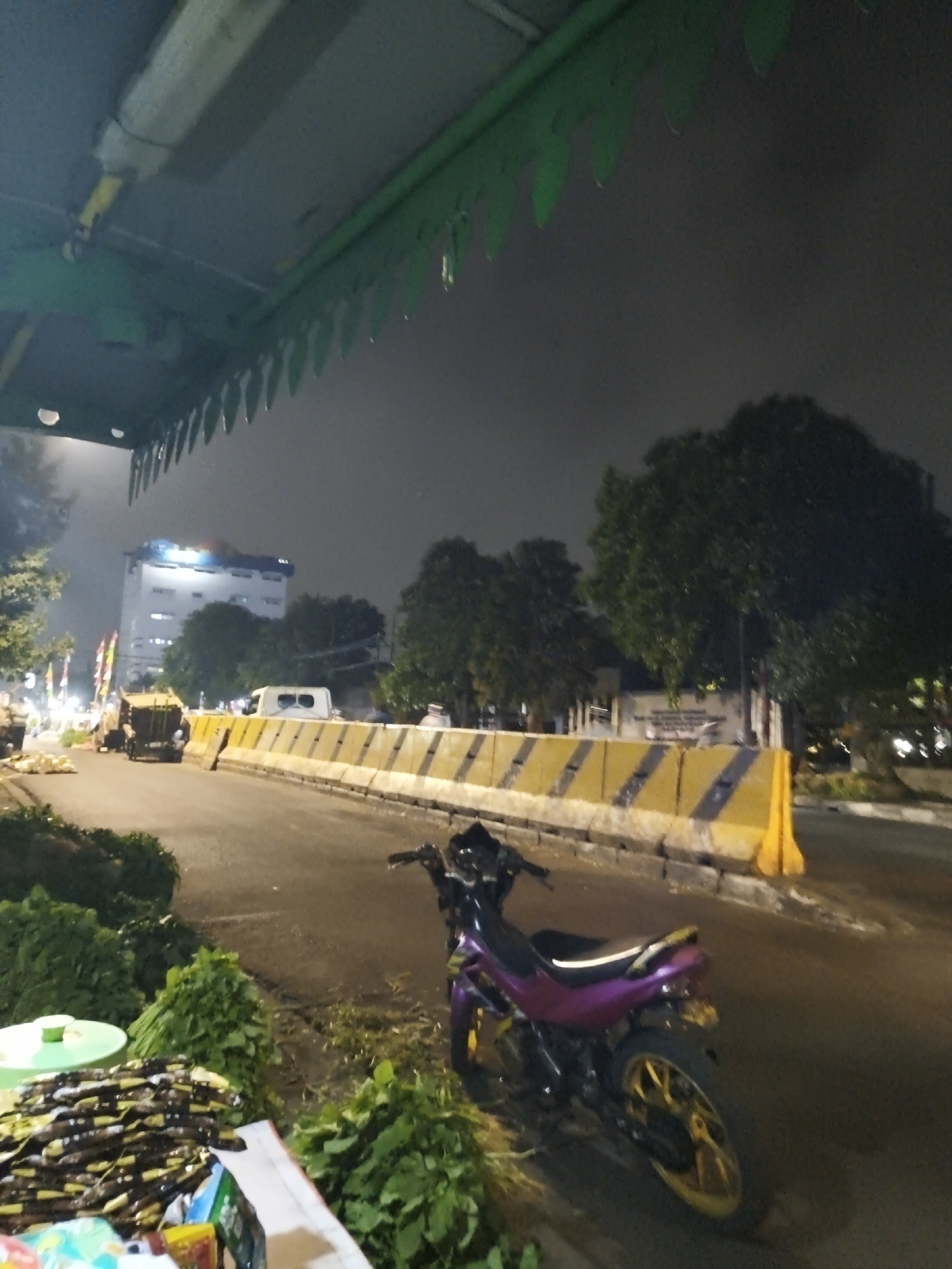 The image depicts a street scene in South Jakarta, Indonesia, at dusk or night. In the foreground, a purple and black motorcycle is parked on the side of a road. To the left of the motorcycle, piles of green leafy vegetables and bundled items, possibly food or merchandise, are displayed on the ground and on a light green table. The road itself is lined with yellow and black concrete barriers. Beyond the barriers, a white van and a garbage truck are visible. In the background, a tall white building with illuminated windows stands against a hazy, dark sky, surrounded by trees. The overall impression is of a roadside stall or market operating in an urban environment after dark.