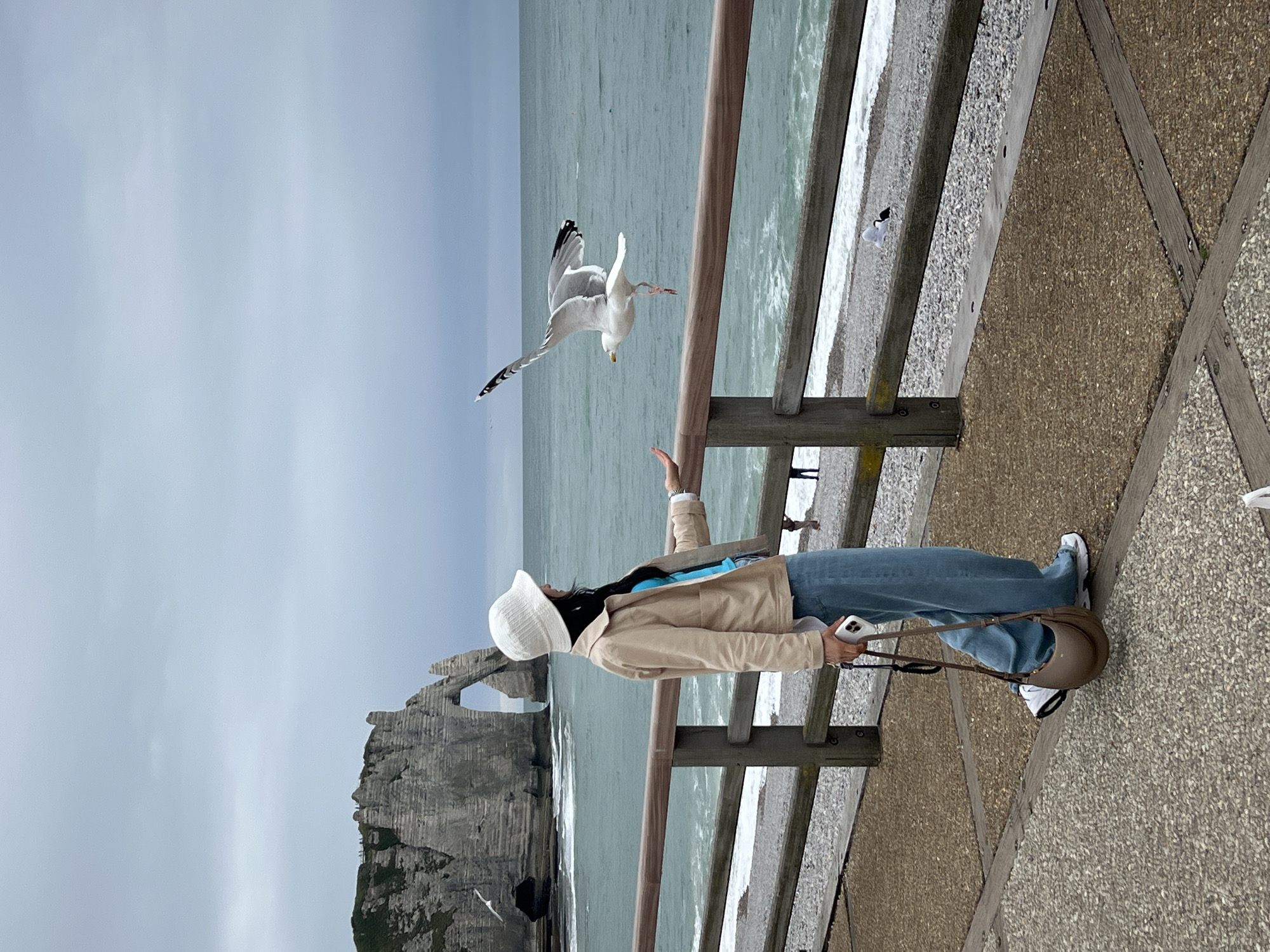 A person wearing a white bucket hat, a beige jacket, and blue jeans stands on a paved walkway by a wooden railing, facing right. They are holding a smartphone in their right hand and a brown shoulder bag hangs from their left arm. A seagull is flying towards the person's outstretched left hand, which is open. In the background, the ocean meets a pebble beach, and dramatic white chalk cliffs with a natural arch formation, characteristic of Étretat, France, are visible under a cloudy sky.
