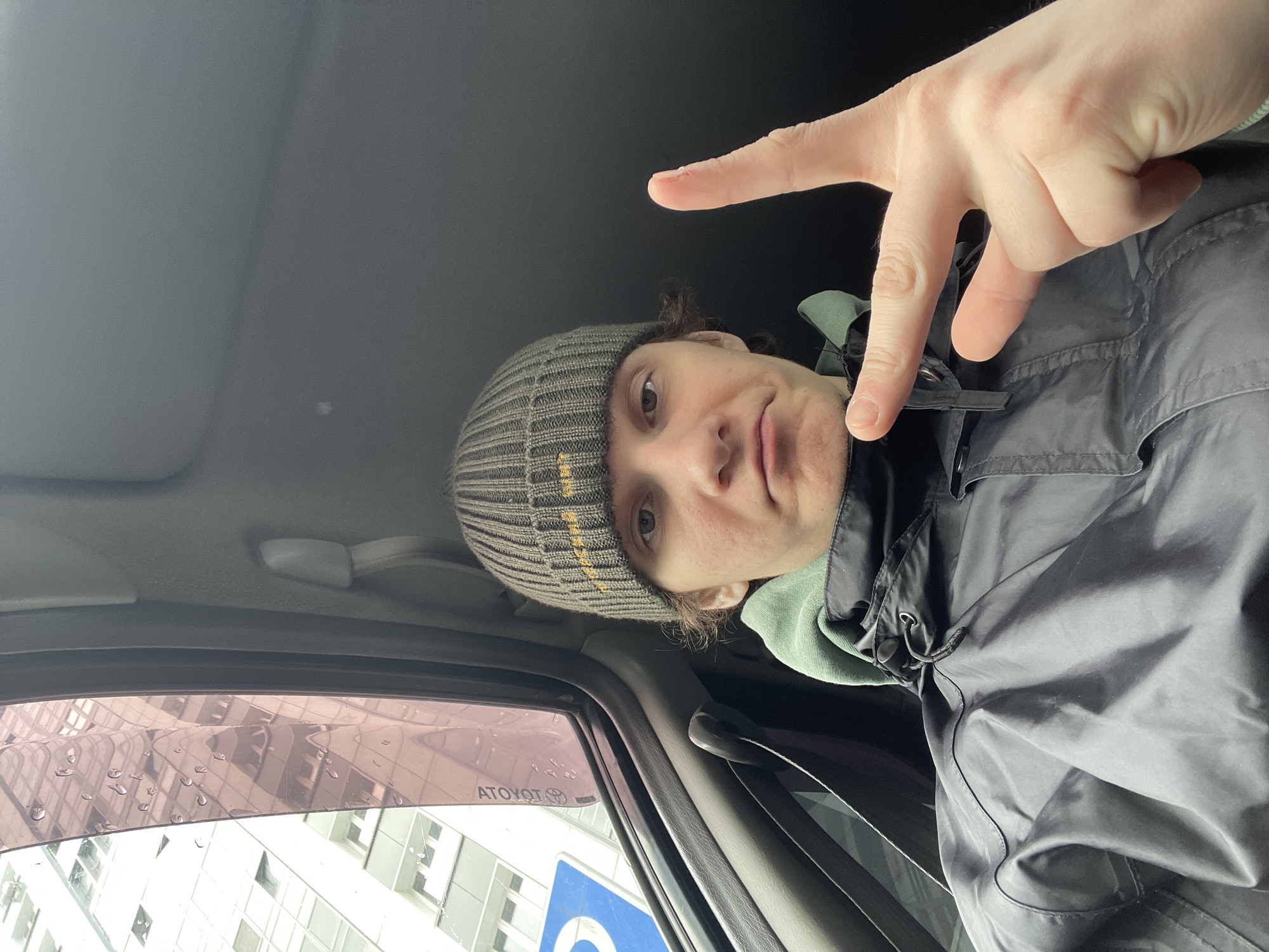 A man is in the driver's seat of a Toyota, wearing a grey beanie with yellow stripes and a dark-colored jacket over a light green hoodie. He is making a hand gesture towards the camera. Outside the car window, a building is visible with a parking sign. The image was likely taken in Krasnoyarsk, Russia.
