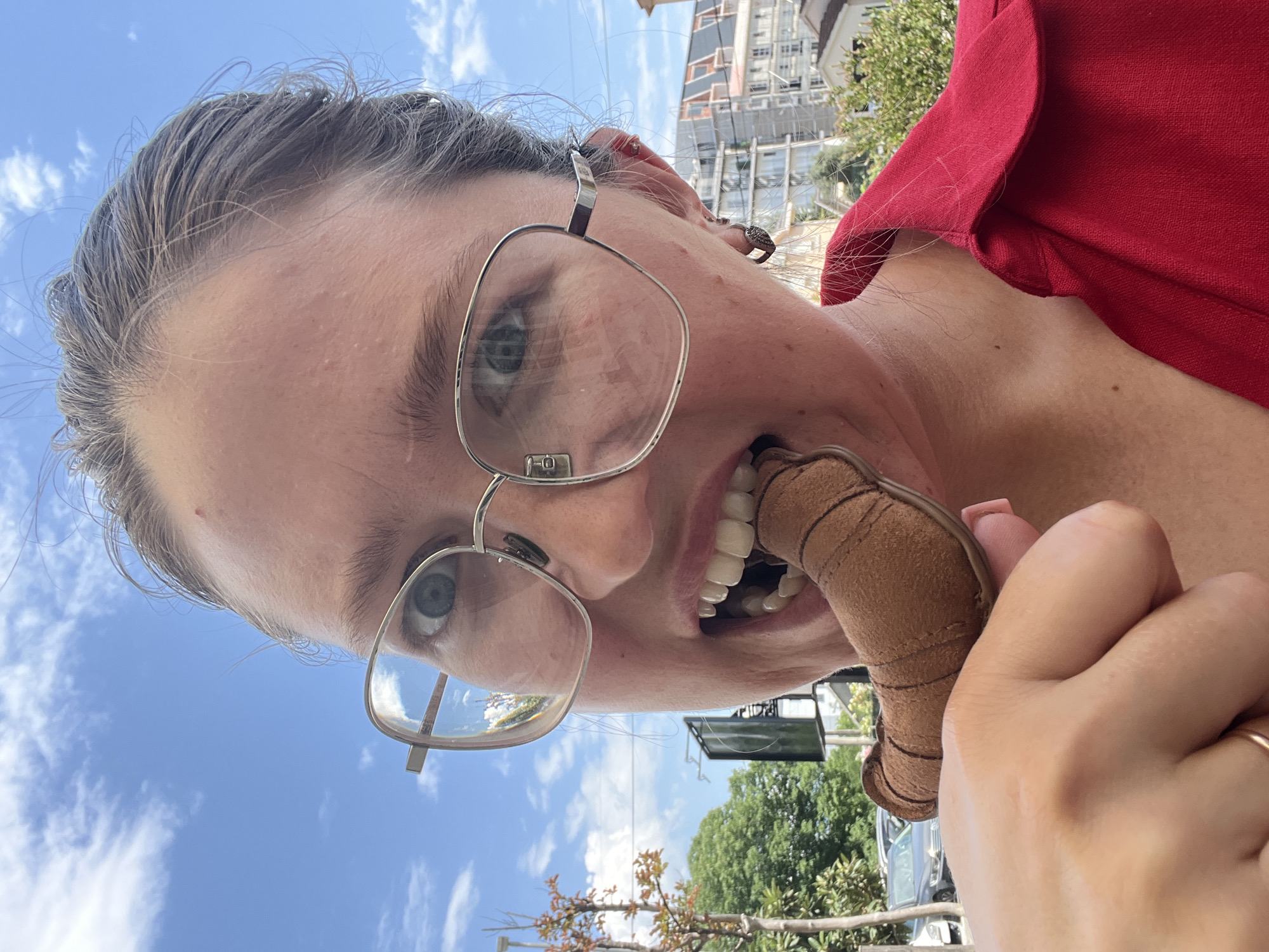 A woman with fair skin and graying hair, wearing glasses and a red blouse, holds a brown suede object in her hand and bites it with her teeth. The picture is taken at a low angle, looking up at her face. The background shows a clear blue sky, trees, and a building under construction. The location appears to be in an urban area, possibly Podgorica, Montenegro, based on the architectural style and construction activity visible in the background.