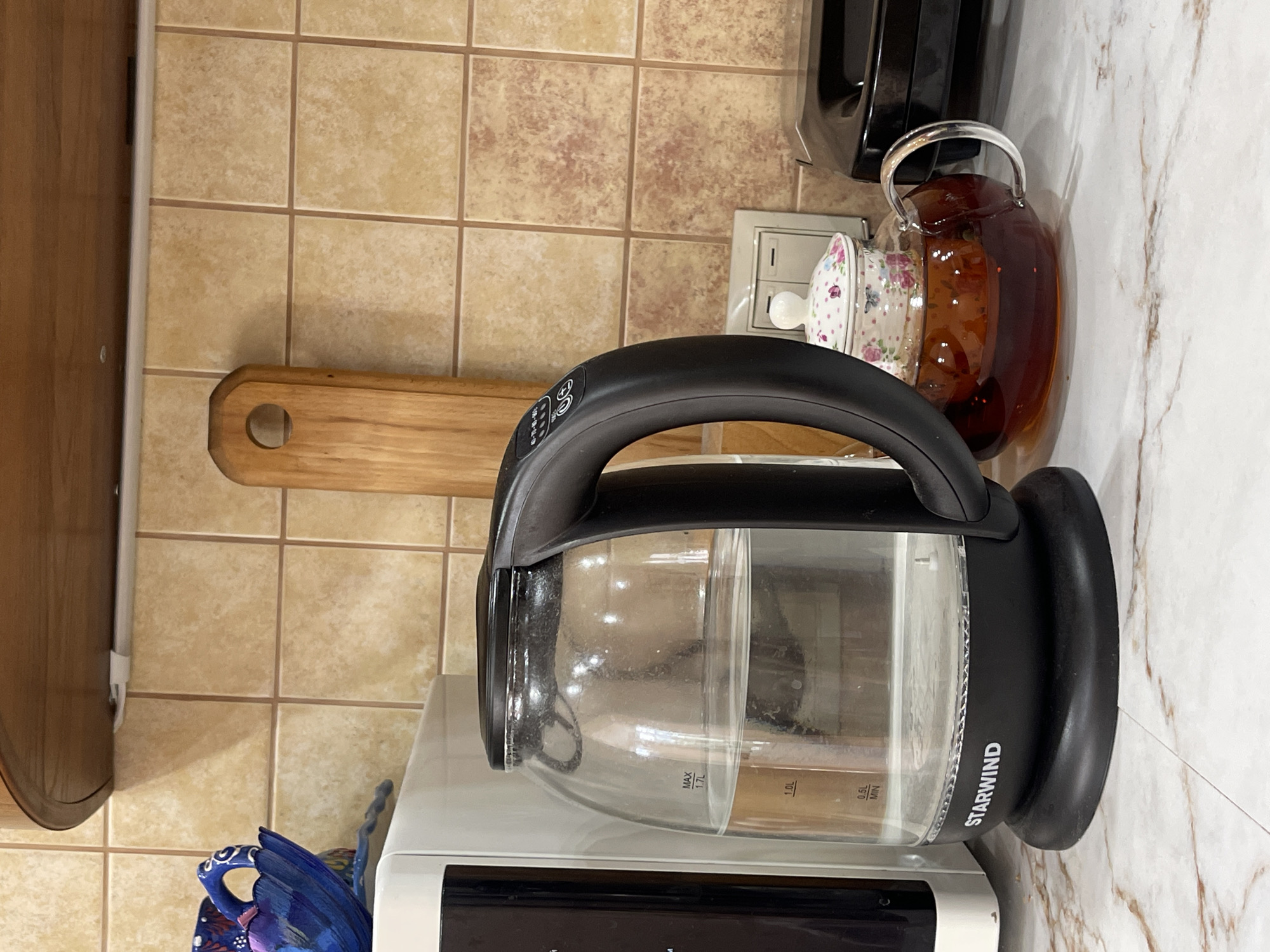 The image shows a kitchen counter with various items. A Starwind electric kettle is prominently displayed, sitting on what appears to be a microwave. Beside the kettle, there is a glass teapot with brown liquid, likely tea. To the left of the kettle is a wooden cutting board hanging on the wall. The wall is tiled with tan, square tiles. Part of a dark-colored coffee maker is visible in the background. The counter itself has a marble pattern. A blue decorative object can be seen in the lower left corner of the frame. The location is Donskoye, Russia.