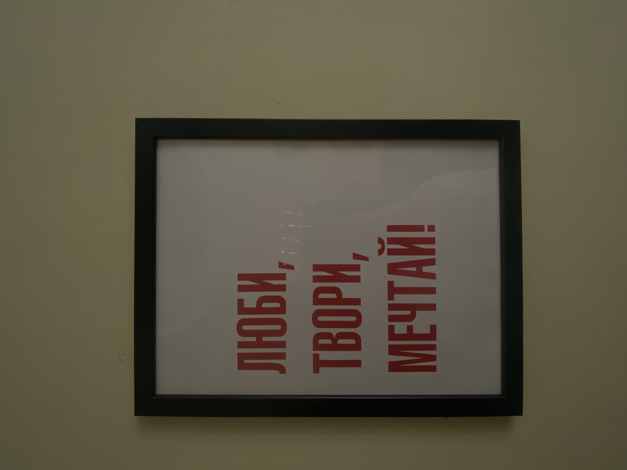 The image displays a framed piece of art or a motivational poster hanging on a textured, light-colored wall, likely indoors in Krasnoyarsk, Russia. The wall is painted in a muted beige or light olive green shade. The artwork itself features a crisp white background enclosed within a simple black frame.
The central element of the poster is bold red text written in Cyrillic. The text is arranged vertically, reading downwards if the frame were upright, but it appears to be rotated ninety degrees counter-clockwise within the frame, so the words read from left to right in an upward direction. The message conveyed is inspirational, comprising three imperative verbs.
The visible text translates as:
"ЛЮБИ," (LYUBI,) meaning "Love,"
"ТВОРИ," (TVORI,) meaning "Create,"
"МЕЧТАЙ!" (MECHTAI!) meaning "Dream!"
Commas separate the first two words, and an exclamation mark follows the final word, emphasizing the motivational sentiment. The lighting in the scene is soft and even, suggesting ambient indoor illumination rather than direct sunlight. There are no people present, and the focus remains entirely on the framed artwork and its immediate, neutral-toned wall background.