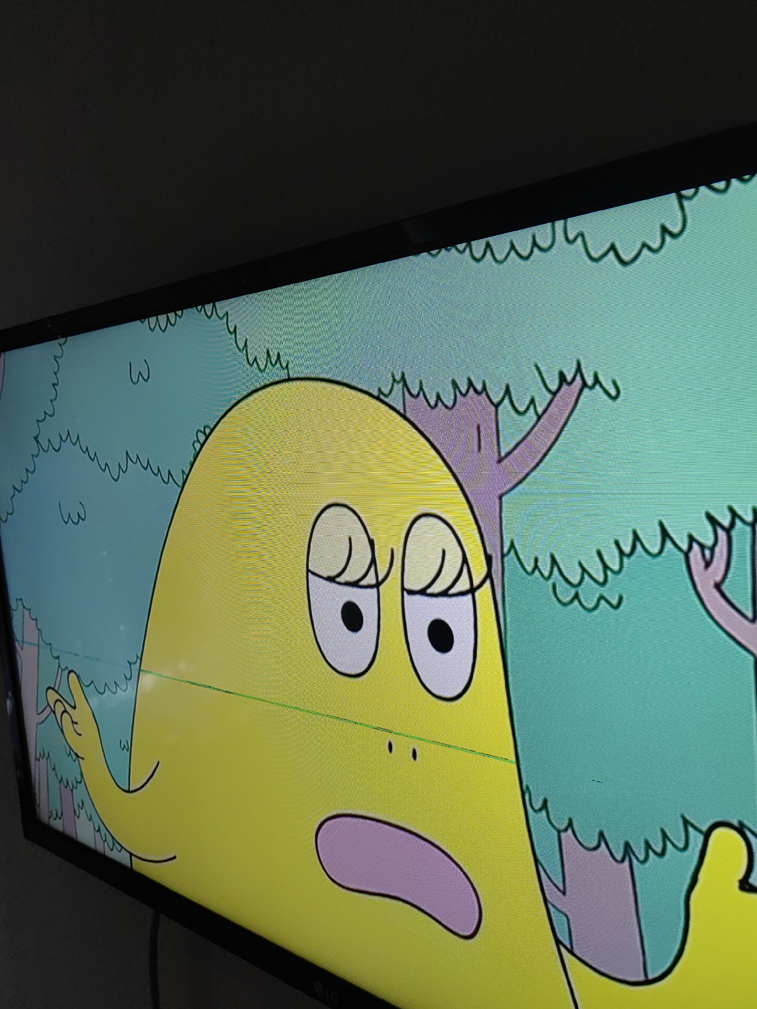 A low-angle, close-up, medium shot captures a cartoon character on a television screen. The character is large, yellow, and appears to have a rounded head with two closed eyes, each outlined with black. One eye is white with a black pupil. A light pink mouth is open, revealing a pink interior. The character's body is primarily visible, and to its left, a hand with three fingers is raised.
The background consists of a light blue sky with light green, wavy lines representing foliage. There are also simplified purple tree trunks and branches with similar wavy green foliage. A thin, bright green horizontal line runs across the screen, partially obscuring the character's mid-section. The television itself is black, and the lower right corner of the screen shows the "LG" logo.
This image is a photograph of a television screen displaying an animated scene. There is no information within the image to suggest a location in Djerovo, Bulgaria.