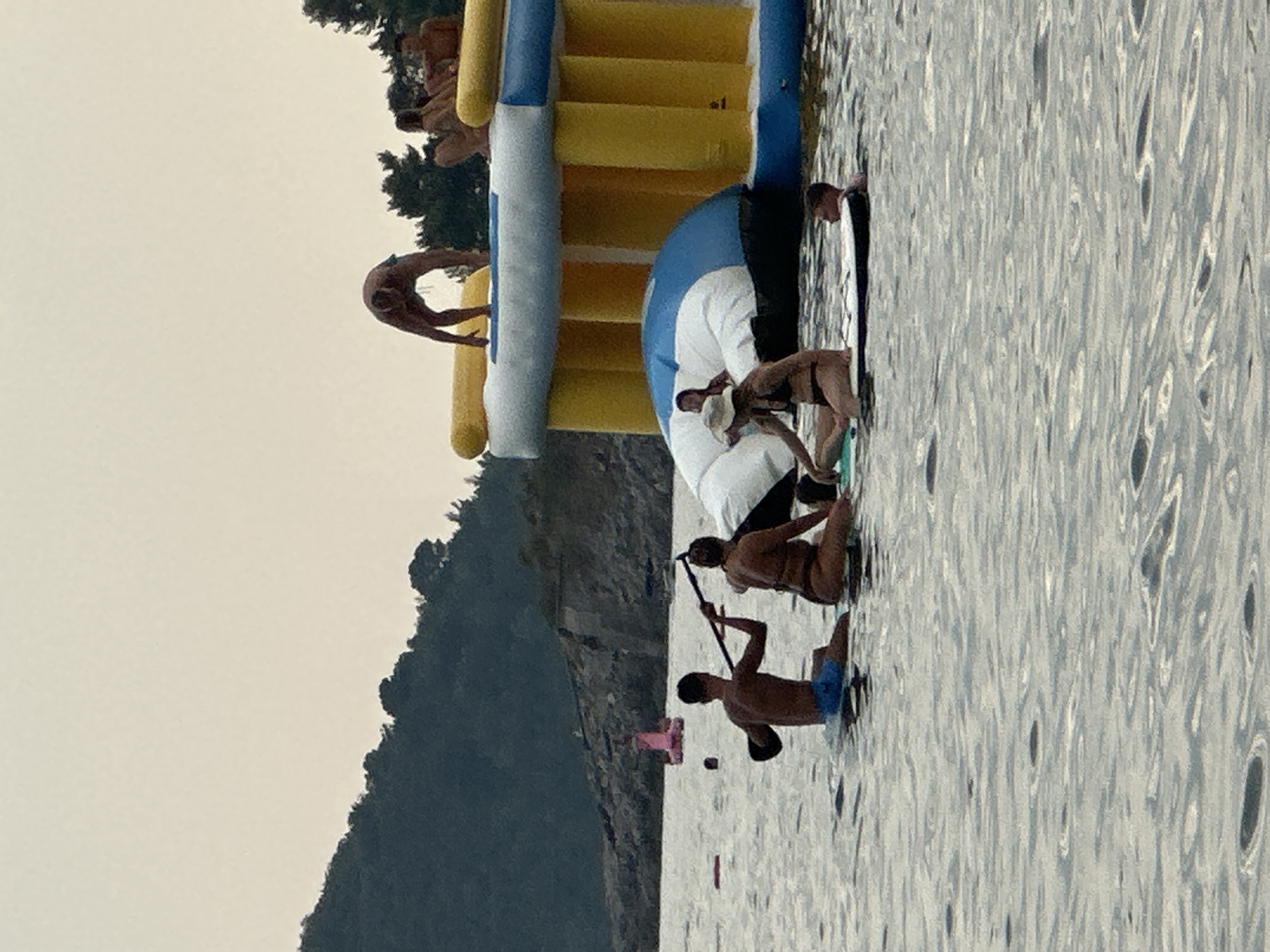 In the waters off Boreti, Montenegro, a group of individuals engages in various water activities. In the foreground, two people are seated on a paddleboard. One, a male wearing blue swim trunks, holds a paddle across the board. The other, a female in a bikini, sits facing away from the viewer, also on the paddleboard. To the right of them, a young male is partially submerged in the water, leaning on what appears to be another paddleboard.
In the background, a large inflatable water park is visible. Several people are on and around the structure. One person is positioned on the top yellow section of the park, appearing to be in motion. Others are seated on the inflatable, observing.
The environment is a body of water with ripples indicating gentle movement. Behind the water park and the people, a hilly, tree-covered coastline rises, typical of the Boreti area. The sky is overcast and light, suggesting a diffused light condition. A pink inflatable object is visible in the middle distance, floating on the water.