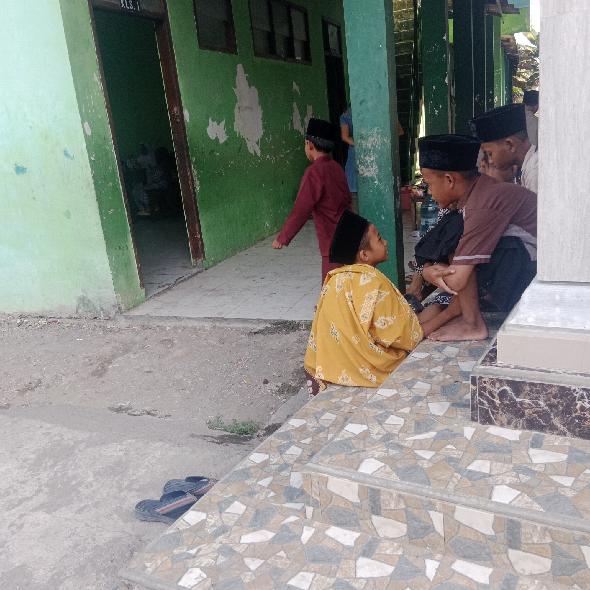 The image depicts an outdoor scene in front of a building with a green, peeling facade, likely a school or community building, in Tegowanu Kulon, Indonesia. The foreground features a tiled staircase with a mosaic pattern of beige, gray, and white tiles. A pair of dark-colored flip-flops with red and white stripes are visible on the ground to the left of the stairs.
Several young boys wearing black peci hats are present. One boy, in the mid-ground, is wearing a yellow, patterned garment and is seated facing to the right, looking upwards. To his right, two other boys are seated. One is wearing a brown shirt and black shorts, and the other is partially visible wearing a white shirt. In the background, a boy in a maroon garment walks away from the camera, and an open doorway reveals a glimpse of individuals inside a room with a sign reading "KLS. 1". The environment appears somewhat worn, with dirt and debris on the ground. The lighting suggests it is daytime.