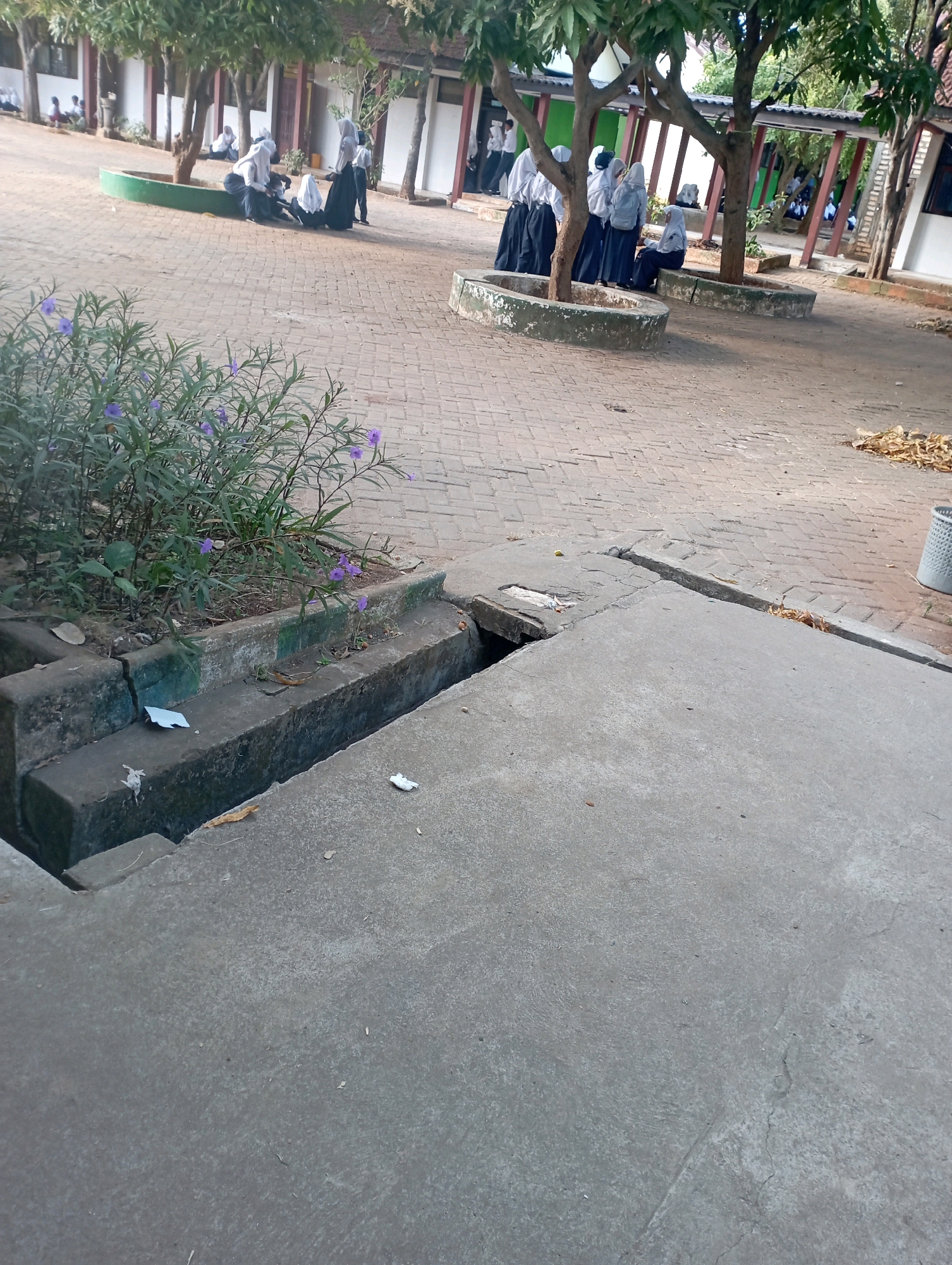 The image captures an outdoor scene in Banyuurip, Indonesia, depicting a group of students in uniform gathered near trees and planters. In the foreground, a concrete drainage ditch is visible, with a patch of purple flowers growing nearby. The ground is paved with brick. Several students, dressed in white tops and dark skirts or trousers, are positioned around circular planters with greenery. Some are standing, while others are seated or leaning against the planters. The environment suggests a schoolyard or communal outdoor space with trees providing shade. The lighting indicates it is daytime.