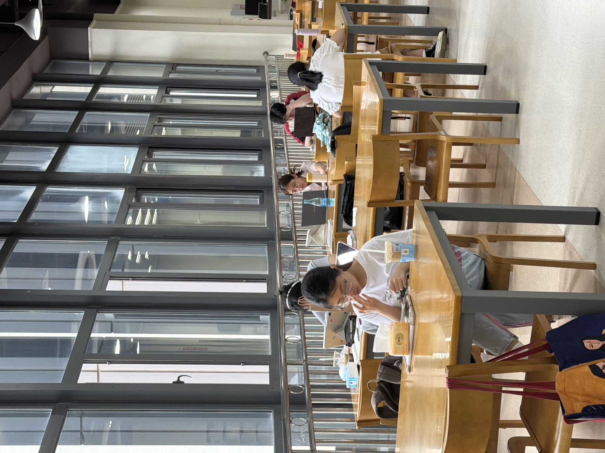 The image depicts a library or study hall setting in Chongqing High-tech Zone, China. Several people are seated at long tables, engaged in studying or working on laptops. The tables are wooden with gray metal frames, and the chairs are wooden. Large windows dominate the left side of the frame, offering natural light. Some individuals have beverages and other items on their tables. The overall atmosphere suggests a quiet, studious environment.