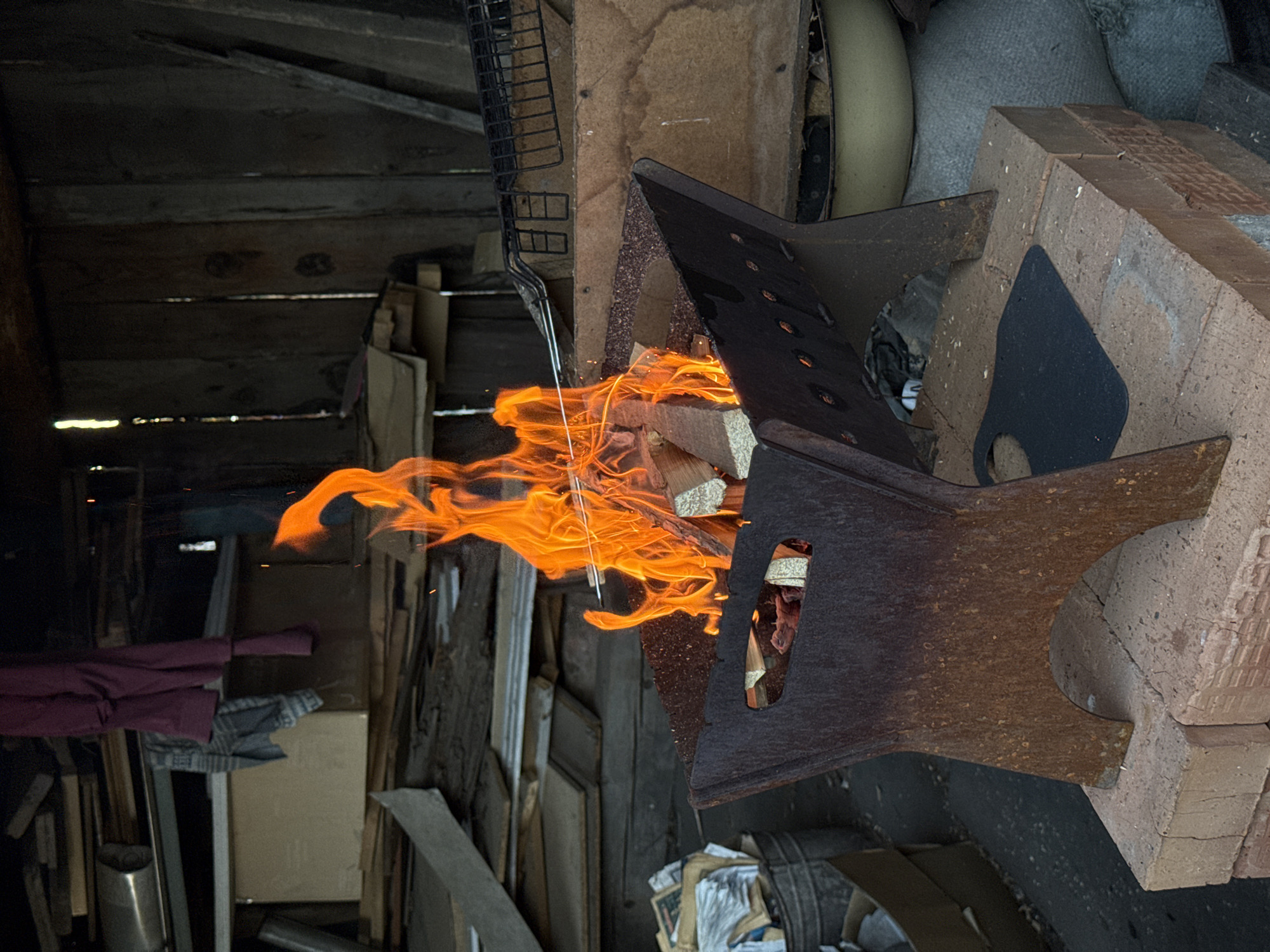 The image shows a fire burning in an indoor setting, possibly a workshop or shed in or near Зыково, Russia. The fire is contained within a dark, rusted metal structure with triangular extensions on top, supported by a brick structure to the right. Flames are visible, consuming what appears to be firewood inside. A metal grill is hanging above and to the left of the fire container. The background includes wooden walls and a cluttered space with various objects, including boxes, fabric, and metallic objects. The scene appears to be illuminated by the fire itself and some natural light filtering in.