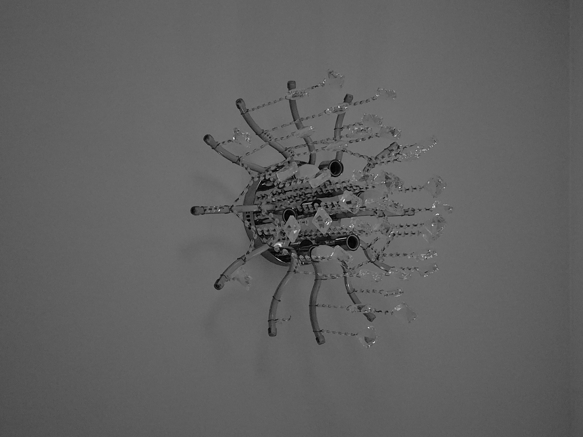 A black and white photograph displays a ceiling-mounted light fixture. The fixture has a central, circular base. Radiating outward are multiple curved arms. Strands of crystal beads hang from these arms. The surrounding area is a flat, uniformly colored ceiling. No people or other objects are visible in the frame. The location of the image is not provided.