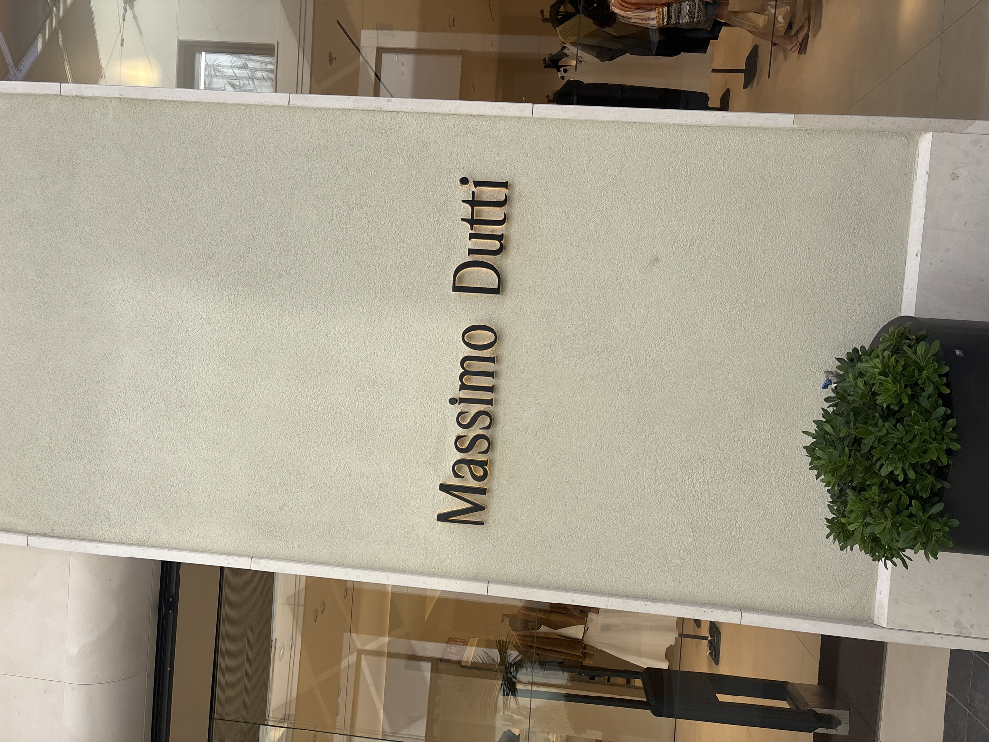 The image depicts the exterior of a Massimo Dutti store, a Spanish fashion retailer, located in the city of Orihuela, Spain. The store's name is prominently displayed in dark, raised lettering against a light beige, textured wall. To the left of the sign, a large glass storefront reveals glimpses of mannequins dressed in clothing, suggesting an indoor retail environment. A dark-colored, slender pillar or structure stands near the entrance. In the foreground, a dark planter with lush green foliage is visible, adding a natural element to the scene. The flooring appears to be a combination of checkered and plain tiled surfaces. The background features reflections in the glass, showing more of the store's interior and potentially other buildings or sky. The lighting suggests it is daytime, possibly overcast given the diffused light. No individuals are clearly identifiable or engaged in significant action, though a person's legs and feet are visible in the lower right portion of the image, indicating pedestrian traffic or customers nearby. The overall impression is of a modern, commercial establishment.