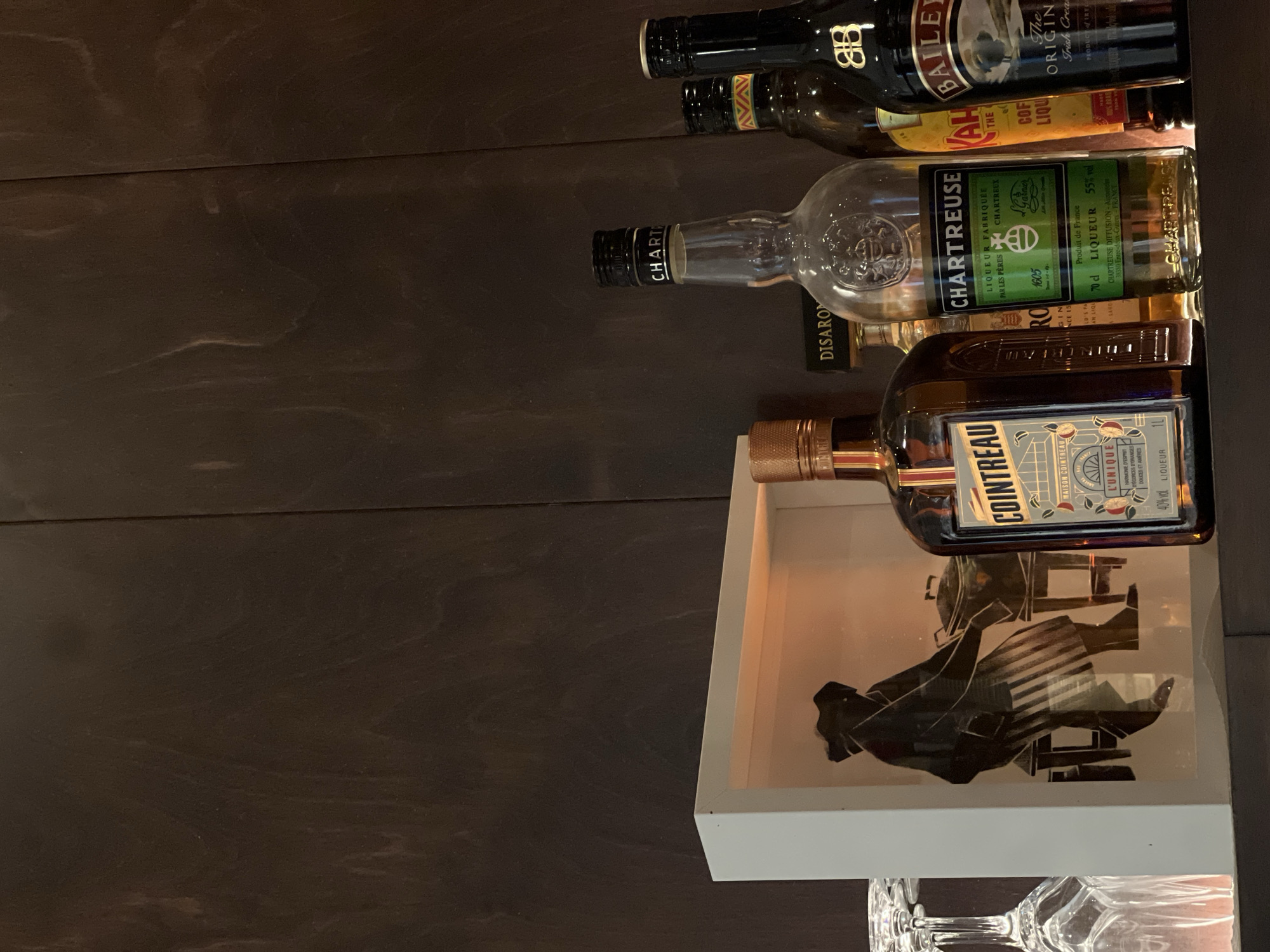 Here is a description of the image:
The image shows a collection of liquor bottles arranged horizontally on a surface, possibly a bar or shelf. The bottles include Cointreau, Disaronno, Chartreuse, Kahlua, and Baileys Irish Cream. Below the Cointreau bottle is a white box with a graphic illustration inside it. Below the white box are glasses. The background is a dark wood surface. The setting is not specified, and there is no information to associate the location with Tel-Aviv, Israel.