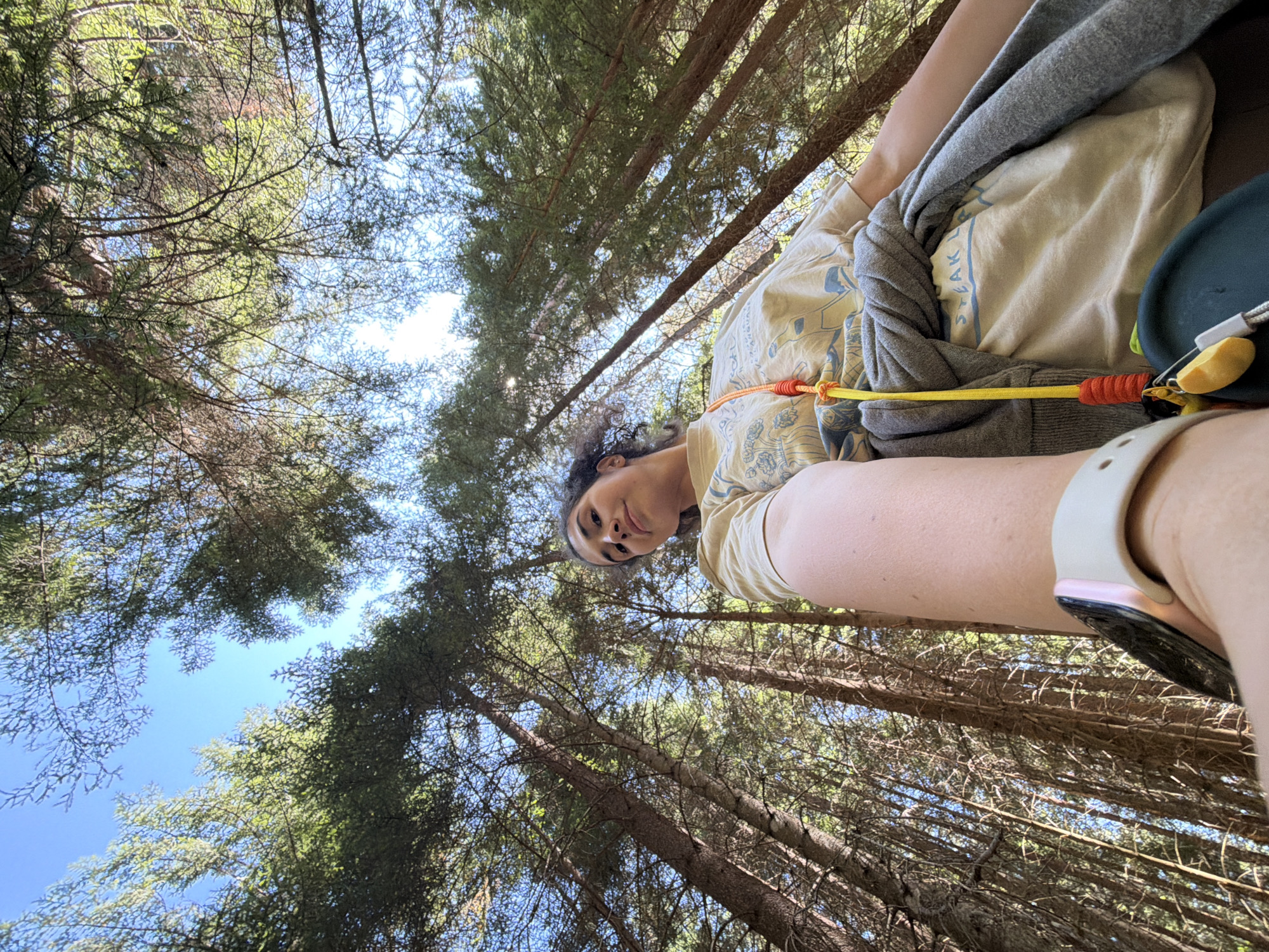 The image shows a low-angle, wide shot of a young woman in a forest, looking up at the camera. She is wearing a light-colored t-shirt with a graphic design and a grey hooded sweatshirt tied around her waist. A colorful lanyard with keys hangs around her neck. She has dark, curly hair and is looking directly at the camera with a slight smile. Her left arm is extended forward, as if taking a selfie. She is wearing a white smartwatch on her left wrist. The background consists of tall evergreen trees, with the blue sky visible through the branches. The scene appears to be in a natural, wooded area, likely a hiking trail or park. No specific landmarks of Sarnitsa, Bulgaria are visible.
