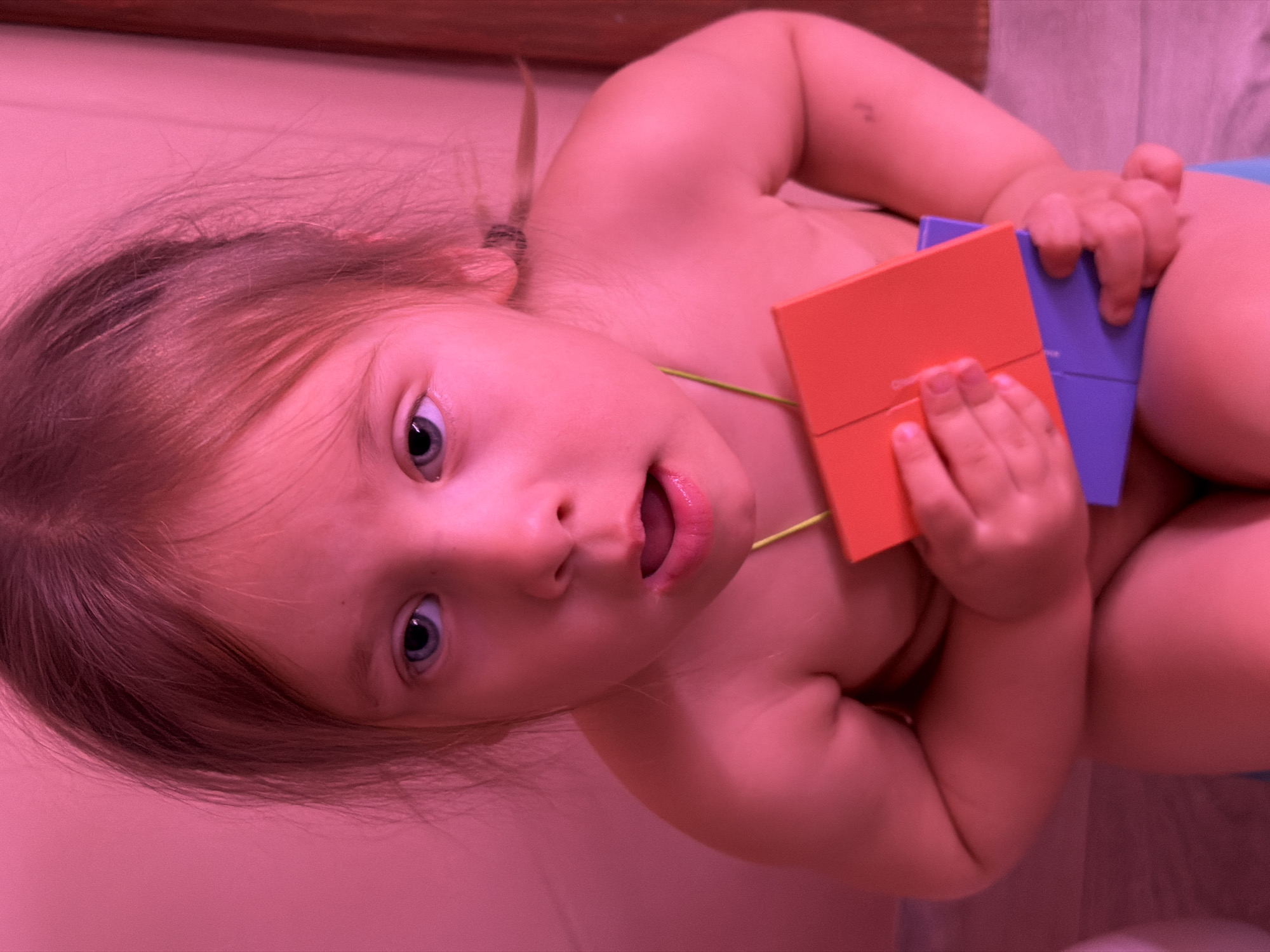 A close-up, slightly angled shot depicts a young child indoors. The child is fair-skinned with light brown hair pulled back into a small ponytail and striking blue eyes. The child's mouth is slightly open, revealing their tongue. In their hands, they hold two square-shaped blocks, one orange and one purple. The scene is bathed in a pinkish-red hue, suggesting the lighting conditions. The background is blurred, but appears to be a plain wall. This image cannot be linked to the city of Chaikovsky, Russia based on visual elements alone.