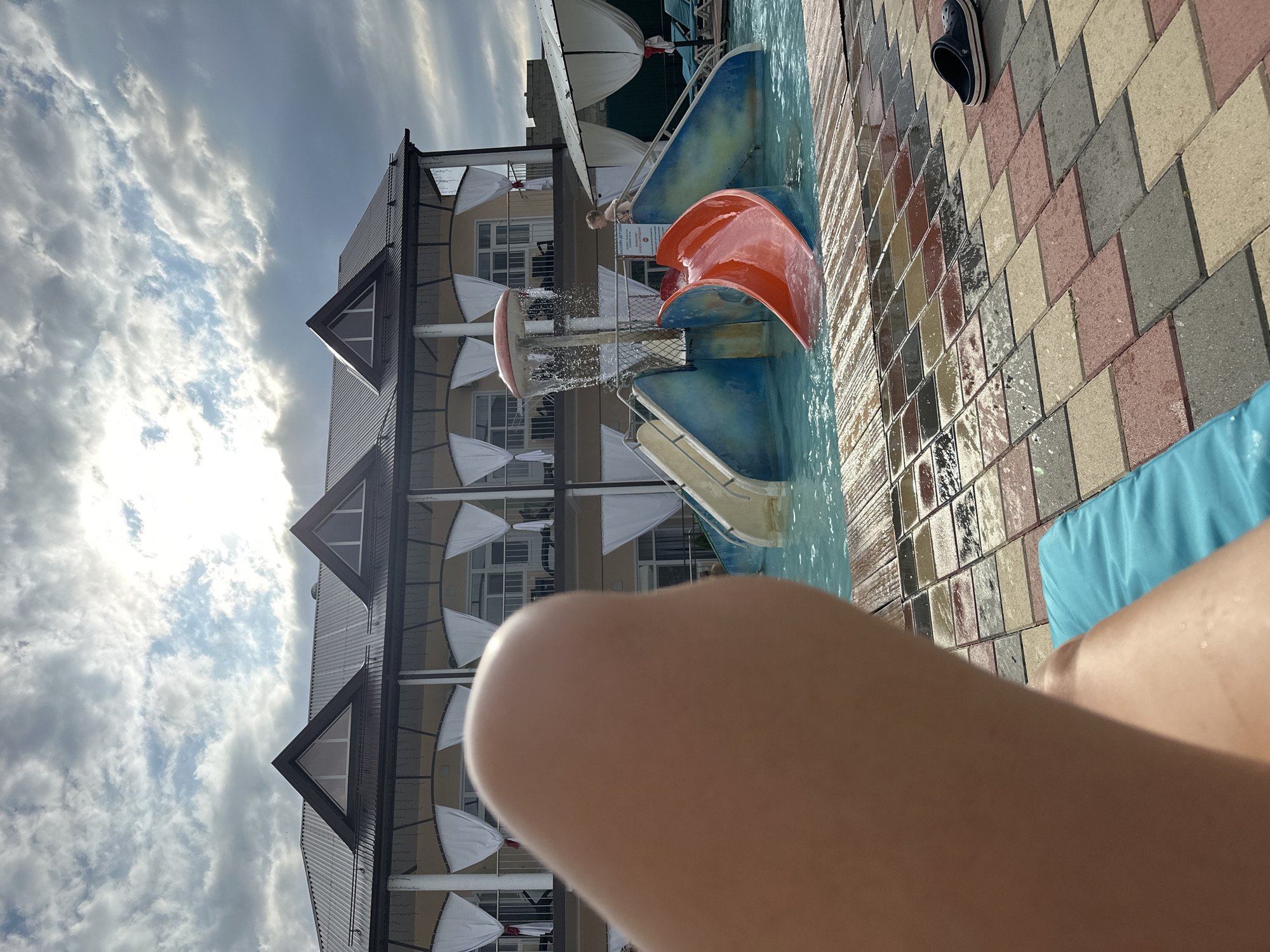 In the city of Yaroslavskaya, Russia, a poolside scene unfolds under a cloudy sky. The foreground is dominated by a blurred close-up of a person's leg and thigh, resting on a turquoise cushion. To the right, a section of the poolside paving is visible, composed of rectangular tiles in various earthy tones. A single navy blue Croc shoe rests on the tiles near the water's edge.
Beyond the paving lies a shallow pool with clear blue water. A children's water slide structure is the central feature of the pool area. The slide is composed of blue and red plastic sections, with a yellow slide on the left and a bright orange slide on the right curving into the water. A mushroom-shaped water feature is positioned above the slides, showering water down.
In the background, a two-story building with a dark, corrugated metal roof stands behind the pool. The building features multiple balconies with white curtains draped across them, suggesting hotel rooms or apartments. The overall atmosphere is one of leisure and relaxation, despite the overcast sky. A young child can be seen standing on the steps leading up to the water slide.