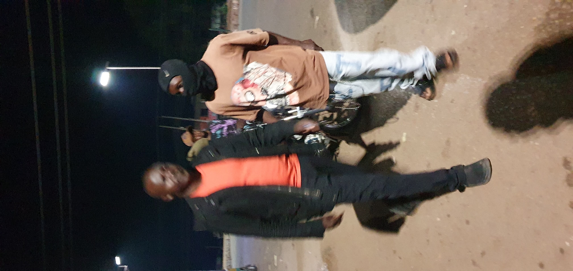 Two men are standing on a dirt road at night, illuminated by artificial lighting. The man on the left is wearing a black jacket over an orange t-shirt and dark pants. He is bald and looking towards the right. The man on the right is wearing a brown t-shirt with a graphic print and light blue jeans, accessorized with a chain. He has a black balaclava covering his face and a black beanie. A motorcycle is partially visible between the two men. In the background, there are streetlights and faint outlines of buildings or structures. The ground is uneven and appears to be a mixture of dirt and asphalt with some puddles. The overall lighting suggests an outdoor urban or semi-urban environment at night.