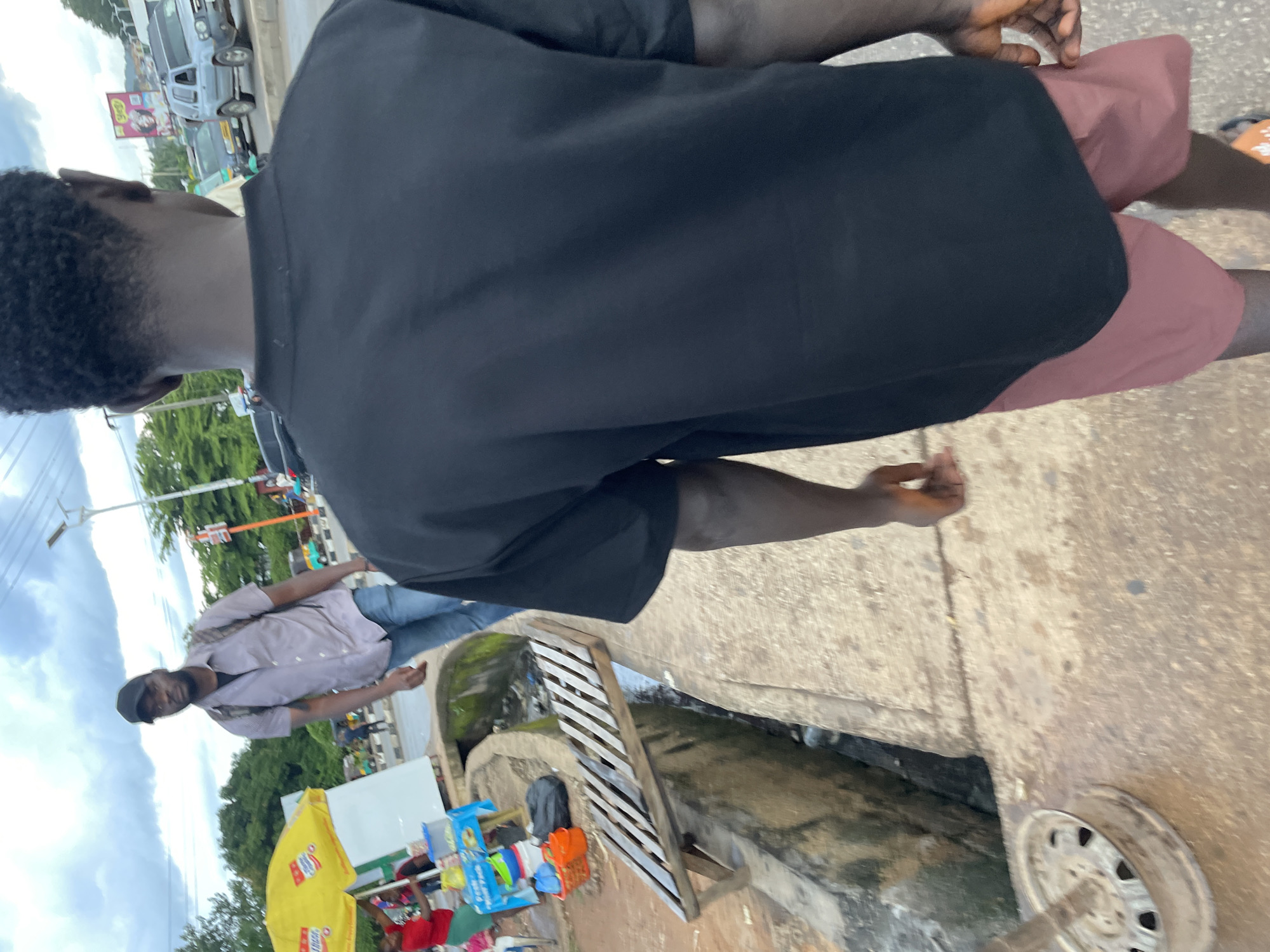 An eye-level, slightly tilted shot captures a bustling street scene in Jos, Nigeria. The foreground is dominated by the back of a young Black man wearing a plain black t-shirt and reddish-brown shorts, his head partially visible. To his left, a man in a light-colored button-down shirt and jeans, wearing a black baseball cap, walks away from the camera, his back to the viewer. He carries a backpack. Behind him, a bright yellow umbrella shades a stall with merchandise. Further back, a ditch runs along the side of the road, its concrete edge lined with debris. A wooden structure, possibly a makeshift bench or shelf, spans part of the ditch. Various vehicles, including a light blue SUV and other cars and motorcycles, are visible on the street, which appears to be a commercial area with shops and vendors. The sky is overcast with dramatic clouds.