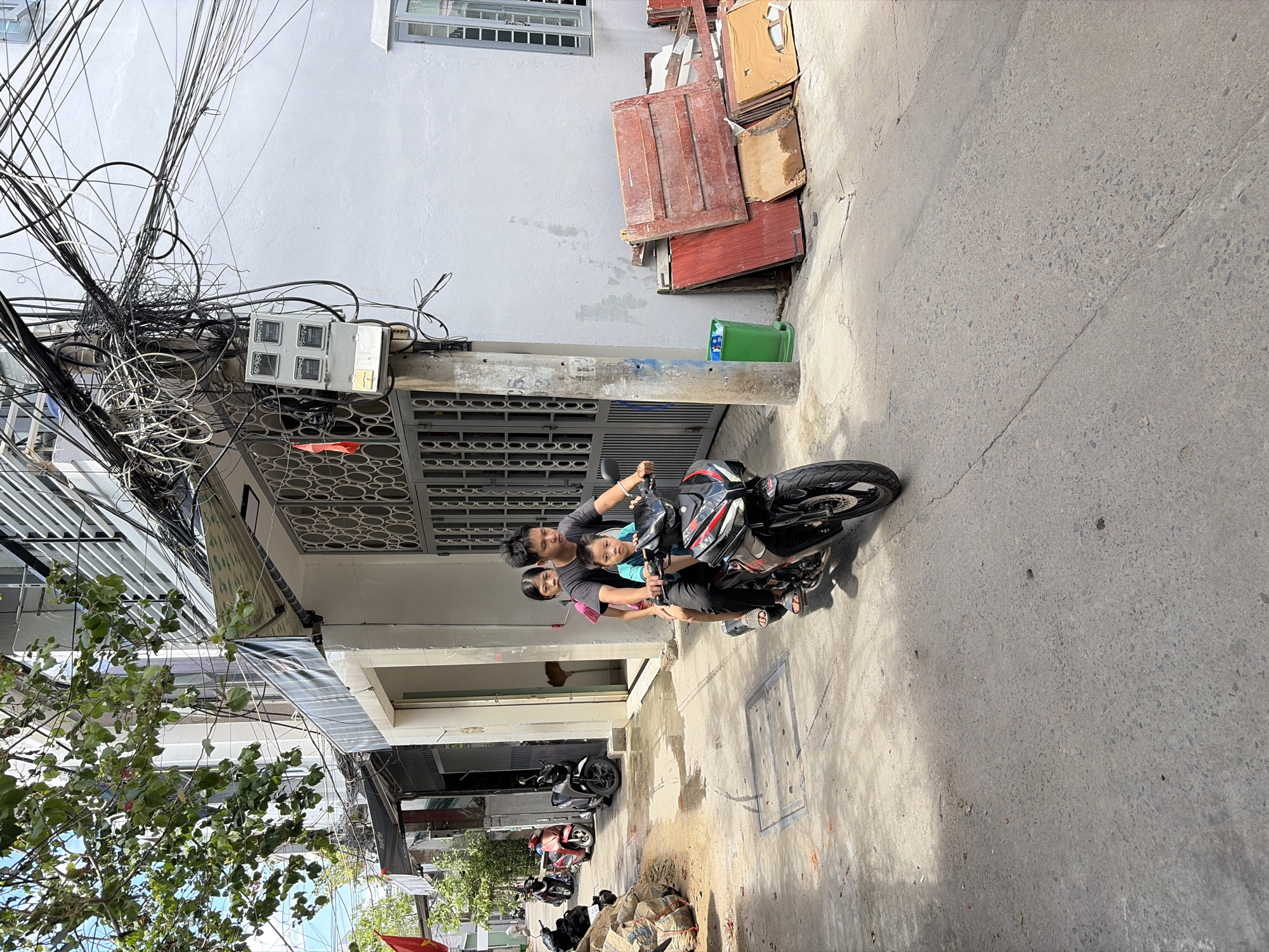 In a narrow urban alleyway in Đà Nẵng, Vietnam, a family of three is riding on a single motorcycle. A man, appearing to be the father, drives the motorcycle, with a young boy seated in front of him and a young girl behind him. They are all looking forward, focused on navigating the street. The alley is lined with buildings displaying a mix of architectural styles, with awnings extending over the sidewalks and a tangle of electrical wires creating a dense network overhead. To the right of the alley, a pile of discarded wooden planks and a green trash bin are visible against a light-colored building. Several other motorcycles are parked along the left side of the alley, suggesting it is a common mode of transportation in the area. The ground is paved with concrete and shows signs of wear and tear, with a manhole cover visible in the foreground. The overall scene depicts a typical day in a densely populated urban environment, with everyday life unfolding on the streets.