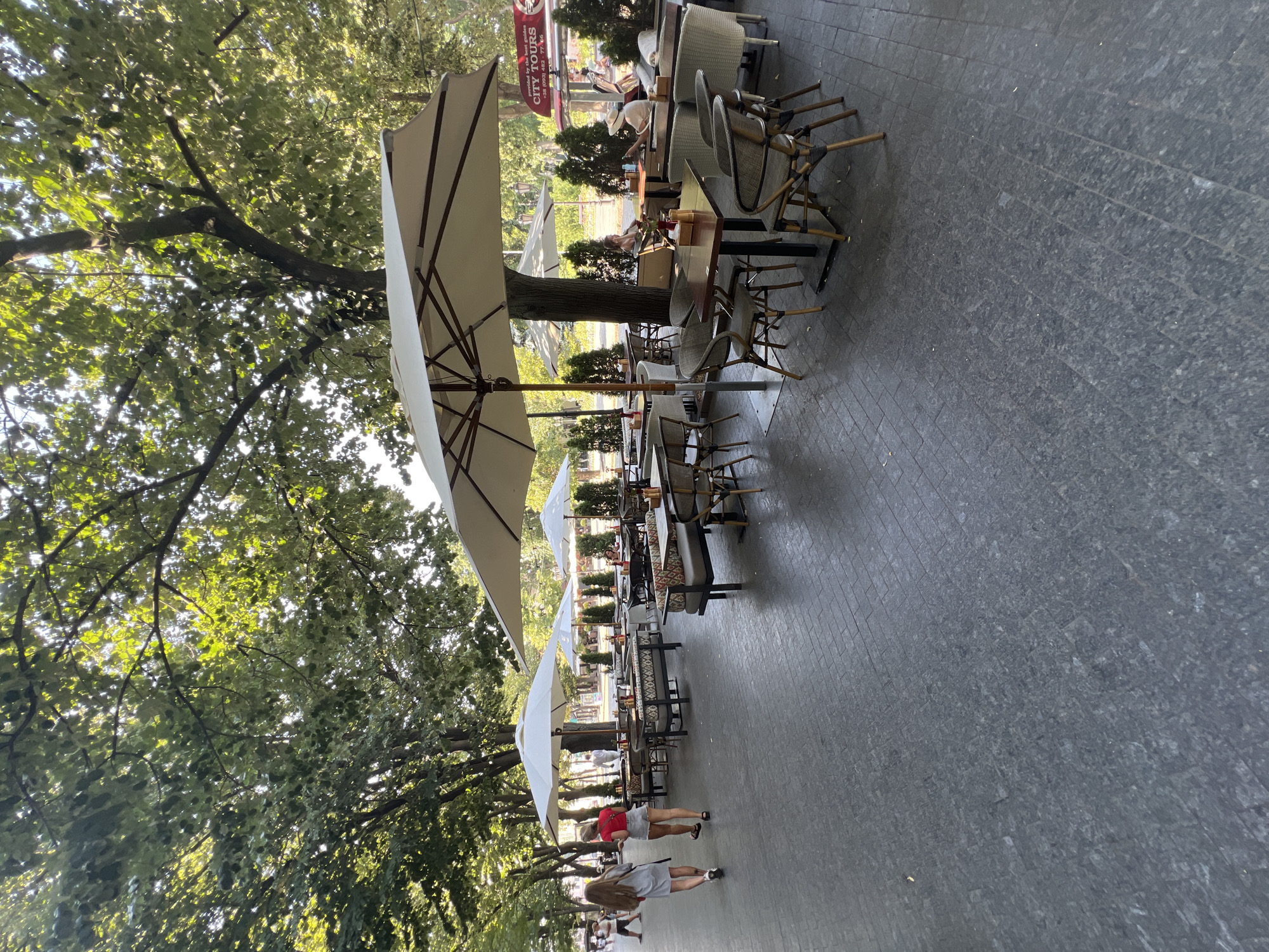 An outdoor seating area of a cafe or restaurant is shown in Odesa, Ukraine. The seating is arranged under large white umbrellas, with tables and chairs on a paved surface. The area is lined with trees, and there is a street visible in the background with a "CITY TOURS" bus. Several people are present, including two women walking away from the camera on the left, and other patrons sitting at tables. The scene appears to be a pleasant, relaxed outdoor dining environment.