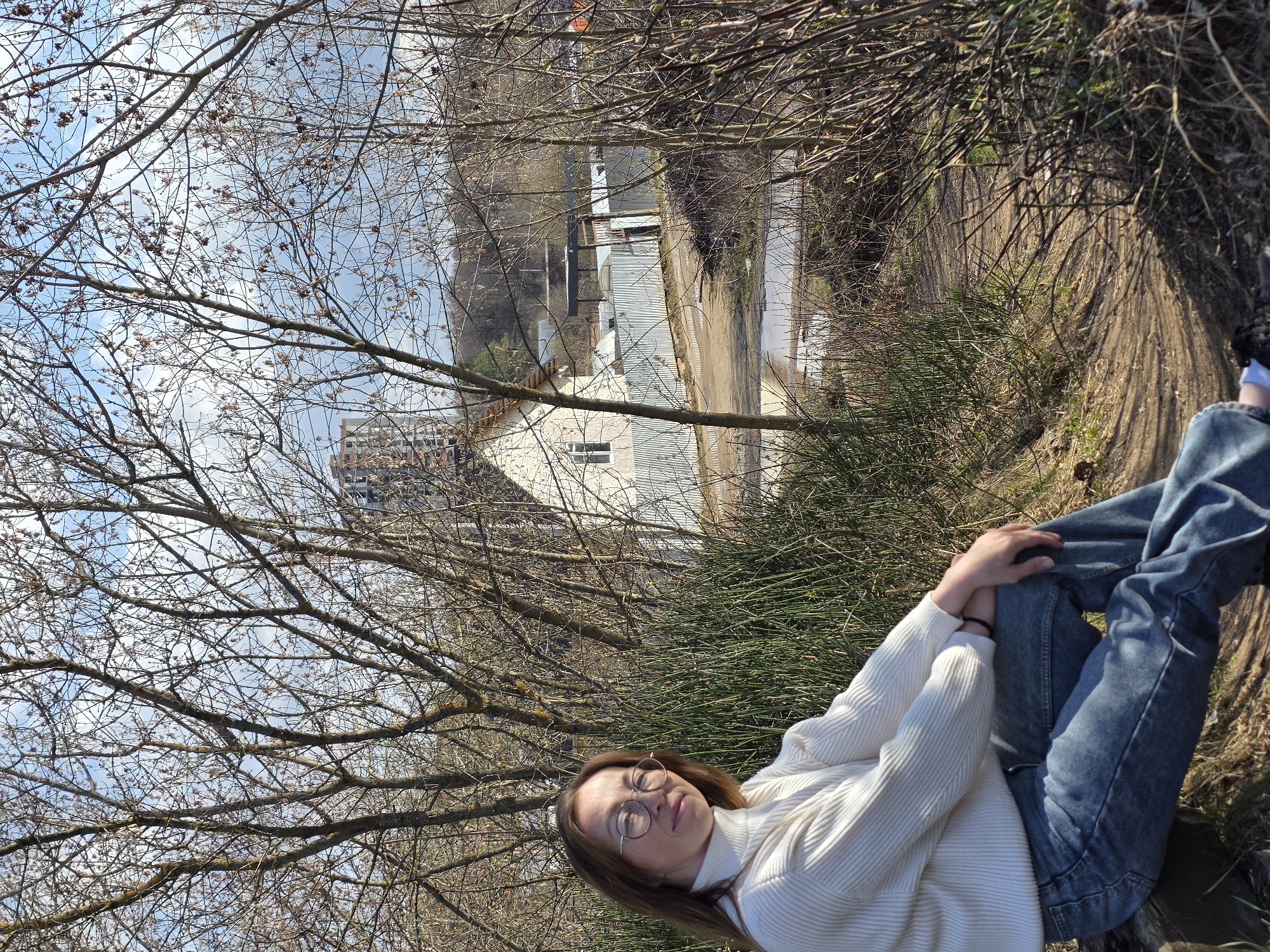 A woman is lying in an outdoor setting in Chaikovsky, Russia. She is wearing a white turtleneck sweater, blue jeans, and round glasses. Her hair is brown and her eyes are closed. She is lying on a dirt path or embankment covered with vegetation. The background includes bare trees, a building with a corrugated metal roof, and a larger building in the distance. The sky is visible through the tree branches.