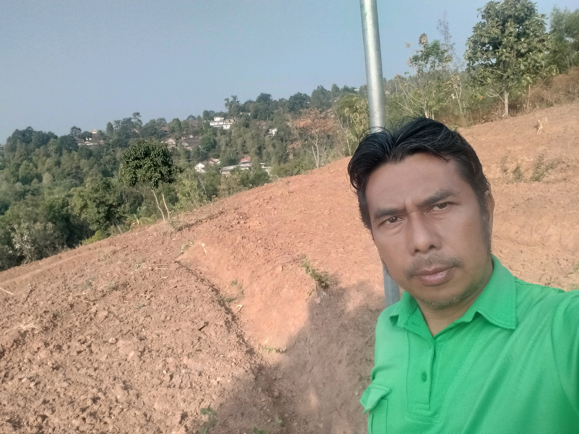 A medium shot captures a man in a green collared shirt taking a selfie on a terraced hillside in Sawahan, Indonesia. The man's face fills the right side of the frame, with his gaze directed towards the camera. Behind him, the landscape unfolds with cultivated terraced fields of reddish-brown soil. In the distance, lush green trees and scattered houses dot the rolling hills under a clear, pale blue sky. A metal pole stands to the left of the man, partially obscuring the background. The overall scene depicts an outdoor environment in a rural or semi-rural Indonesian setting.