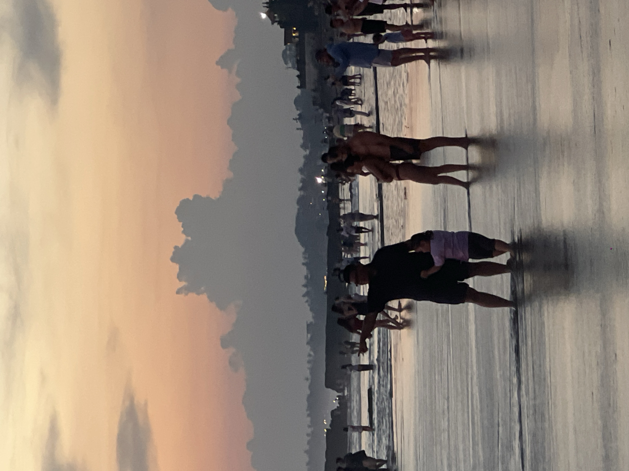 This image depicts a crowded beach at dusk, likely in Canggu, Indonesia, given the coastal setting and the presence of multiple people walking along the shoreline. The sky is a gradient of pink and gray, suggesting sunset or sunrise. Many individuals are visible, some in swimsuits, others in casual wear, engaged in activities like walking, standing, and interacting with each other. A man is holding a small child. The beach appears wet, reflecting the sky and figures, creating long, distorted shadows. Distant lights suggest some development along the coast, such as buildings or streetlights.