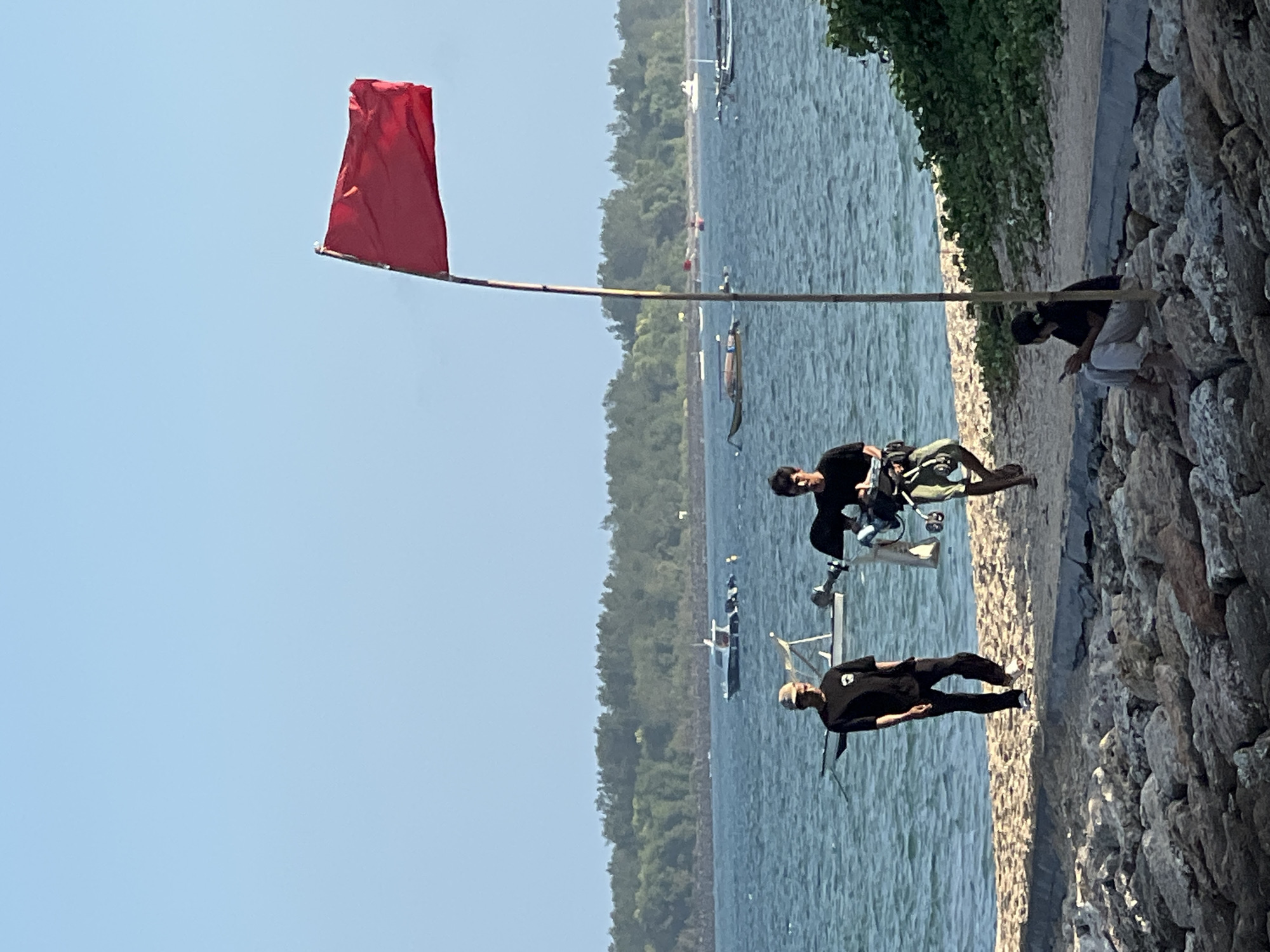 The image captures a beach scene on a sunny day in Denpasar, Indonesia, likely at a location like Sanur Beach given the calm waters and presence of traditional Jukung boats. In the foreground, a rocky shoreline borders a sandy beach. A red flag flies prominently on a bamboo pole, signaling caution or prohibition in the water. Two young men are walking on the beach, one carrying equipment. Further down the beach, a young woman is seated on the rocks, looking at her phone. The sea is visible with several boats anchored or moving in the distance, and a lush green treeline forms the backdrop along the coast. The sky is clear and blue.