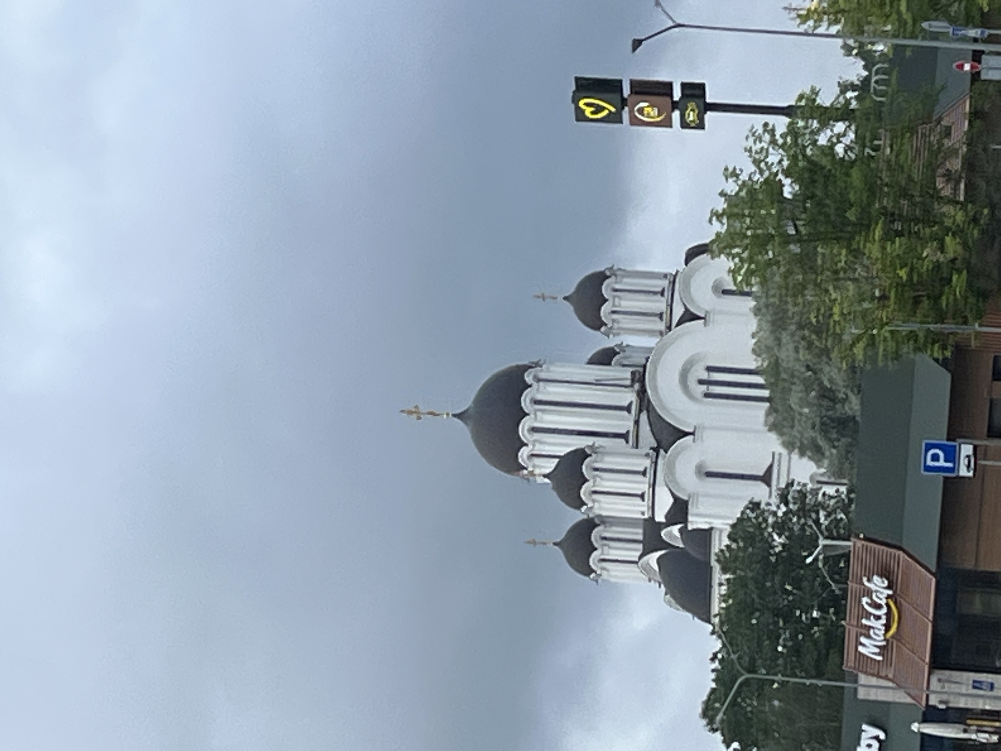 The image captures a cityscape with a prominent white Orthodox church featuring black domes and crosses against a cloudy sky. In the foreground, a McDonald's restaurant sign with "MakCafe" is visible. To the right of the church, a tall sign displays a yellow heart and a circular logo, likely associated with McDonald's. Trees and streetlights are interspersed throughout the scene, suggesting an urban environment. The location is identified as Minsk, Belarus.