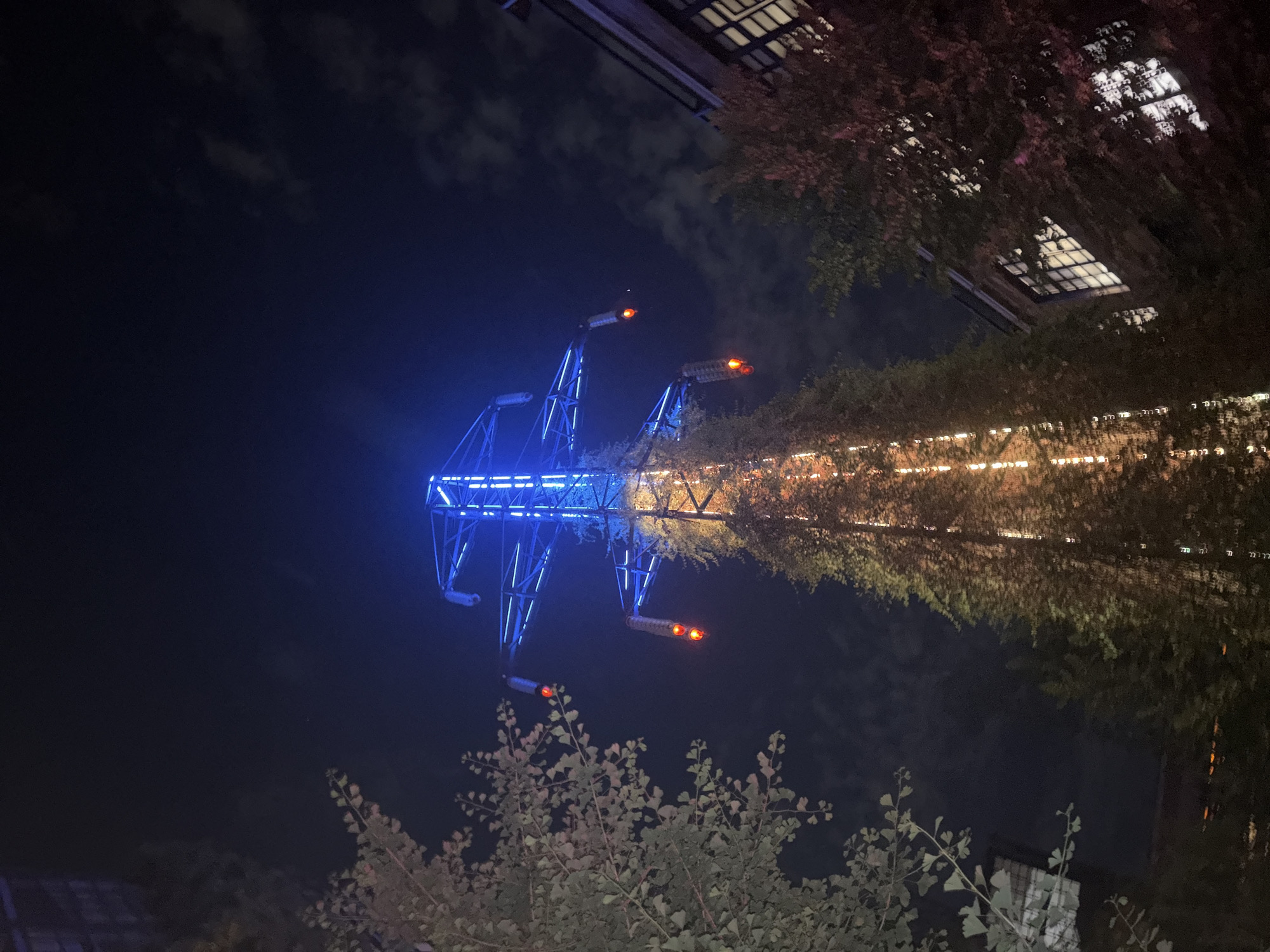 The image shows a nighttime view of a repurposed electricity pylon in Tbilisi, Georgia. The pylon's upper structure is illuminated with blue LED lights, while the lower portion is covered in dense green foliage, also illuminated by warm white lights. Two large, red, cylindrical lights hang from the pylon's arms, resembling traffic signals. To the left and right of the pylon, trees with various leaf colors, some appearing to be ginkgo leaves, frame the structure. In the background, parts of buildings are visible, with windows illuminated from within. The sky is dark, with scattered clouds. The overall scene suggests an urban art installation or a creatively repurposed industrial element within the city.