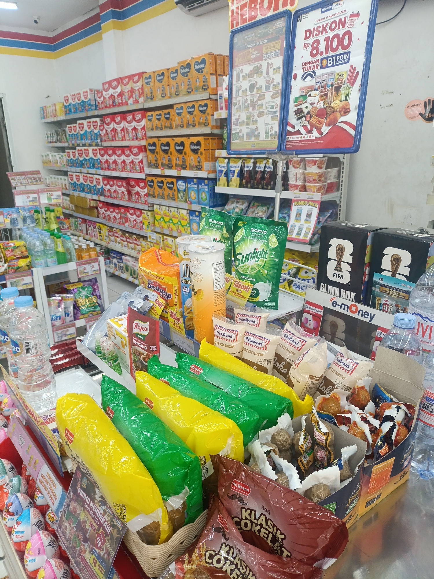 The image depicts the interior of a minimarket, likely Indomaret based on the branding visible on signage and products. The scene is brightly lit, with fluorescent lights overhead, illuminating rows of shelves stocked with various consumer goods. Prominently displayed are packaged food items, beverages, and cleaning supplies. In the foreground, there are several baskets and displays containing snacks, including individually wrapped items and larger bags. One display features yellow and green plastic-wrapped bread or pastries. Another basket holds what appear to be fried snacks in individual packaging. In the background, shelves are laden with boxed milk powder, bottled water, and detergents. Two large blue promotional posters are attached to stands in the middle ground. One poster advertises a discount of Rp 8,100 with 81 loyalty points, featuring images of food items and a FIFA World Cup theme. The other poster displays a variety of products with prices and promotional text. The minimarket is organized and appears to be well-stocked, suggesting a typical retail environment in Indonesia. There are no people visible in the image. The overall impression is that of a busy commercial space offering a wide range of everyday necessities.