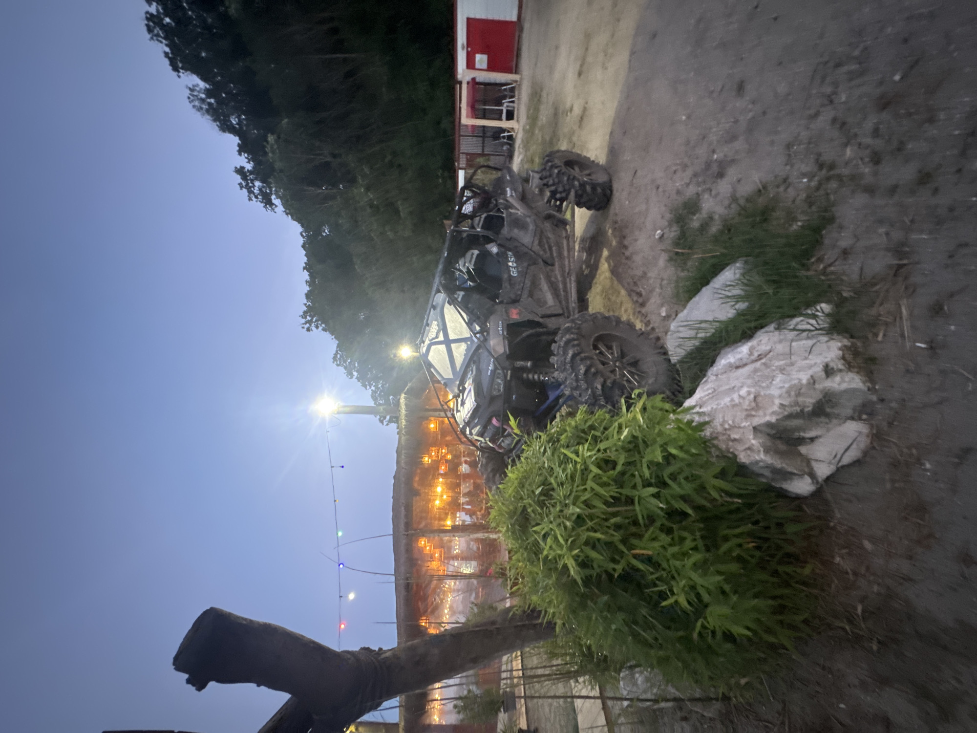 This image depicts an outdoor scene at dusk, likely near a restaurant or beach bar, situated in the city of Varna, Bulgaria. The foreground is dominated by a muddy, all-terrain vehicle (ATV) parked on a dirt and gravel surface. Lush green foliage and large, light-colored rocks are positioned to the left of the ATV. In the mid-ground, a thatched-roof structure, possibly an open-air dining area, is illuminated by numerous warm-toned lights, with clear plastic sheeting enclosing some sections. A string of colorful lights stretches across the top of this structure and into the background. The background features a dense, dark green forest or wooded area. The sky is a pale blue, transitioning to a deeper shade of blue as it rises, indicating twilight. A tall, weathered wooden post stands to the left of the frame. Red and white buildings are visible in the far right background. No people are clearly identifiable in the image.