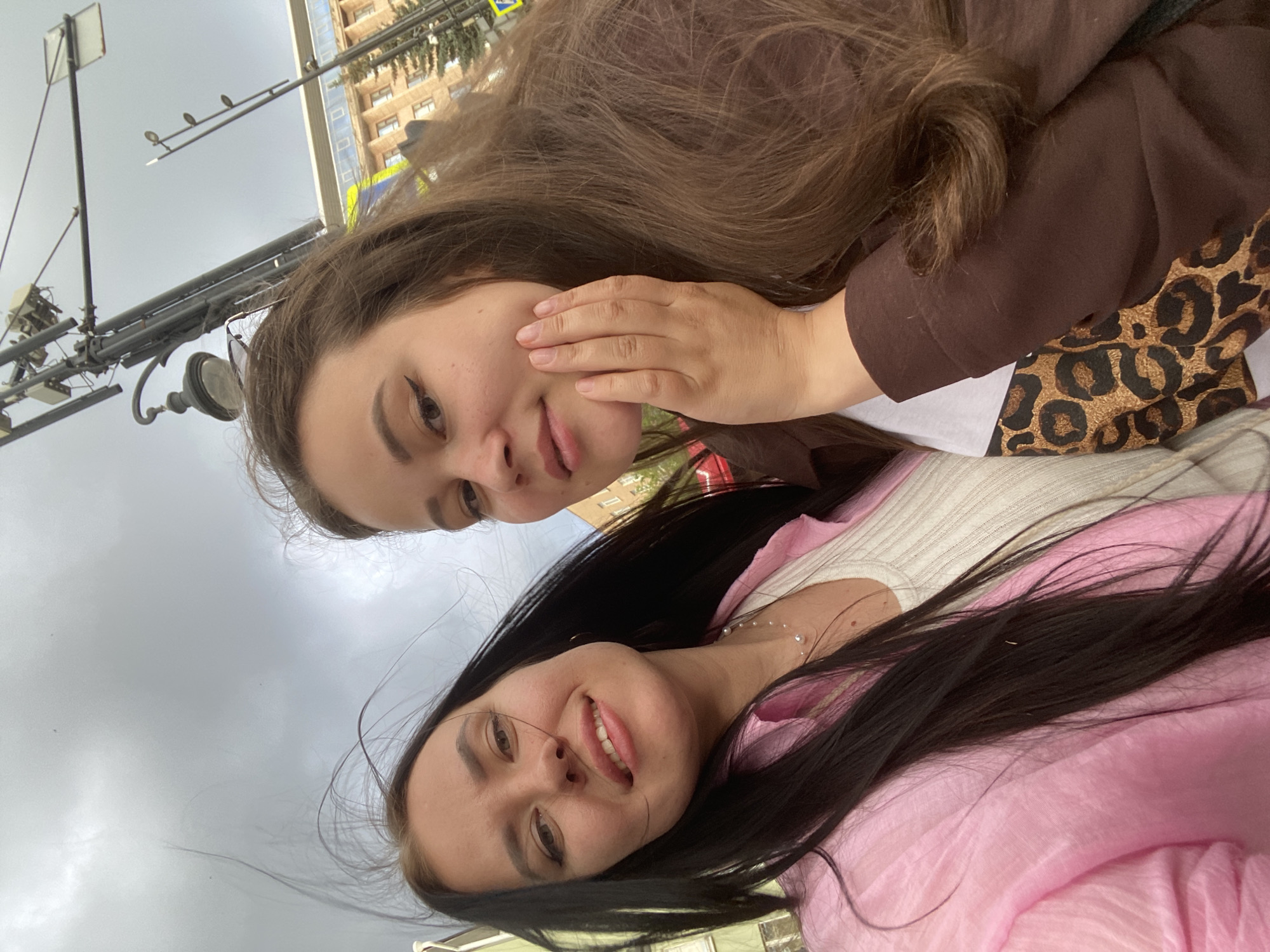 Two young women are captured in a selfie taken outdoors, with a cloudy sky overhead. The woman on the left has long, dark hair and is wearing a light pink collared shirt over a white ribbed top and a pearl necklace. The woman on the right has long, brown hair and is wearing a brown jacket over a leopard-print shirt. She is touching her cheek with her right hand. In the background, there are streetlights, traffic signals, and a building with a light-colored facade. The environment appears to be an urban setting, possibly a city street in Perm, Russia, given the visible street infrastructure and architectural style. The overall mood is casual and friendly.