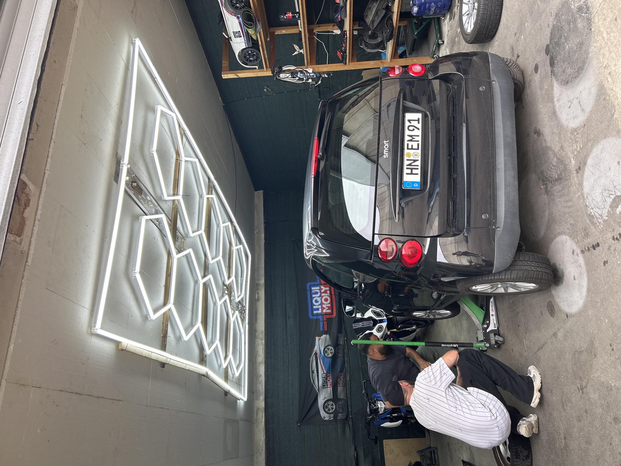 The image depicts a garage with a black Smart car being worked on by two men. One man, wearing a black t-shirt and a black baseball cap, is crouching near the rear wheel, appearing to be assisting with the repair. Another man, wearing a white and black striped t-shirt and black pants, is also crouched down, facing away from the camera, near the car's jack. A green hydraulic jack is positioned under the car's chassis. To the left of the Smart car, a black motorcycle is visible. In the background, a banner advertising "LIQUI MOLY" is hung on a dark green wall, featuring an image of a silver car. To the right of the Smart car, a wooden shelf unit is stocked with various items, including what appears to be a toy car, tools, and bottles. The garage floor is concrete, with oil stains and a detached car tire. Overhead, a hexagonal-shaped lighting fixture with neon tubes illuminates the space. The ceiling appears to be made of concrete or a similar material. The overall environment suggests a car repair or maintenance setting.