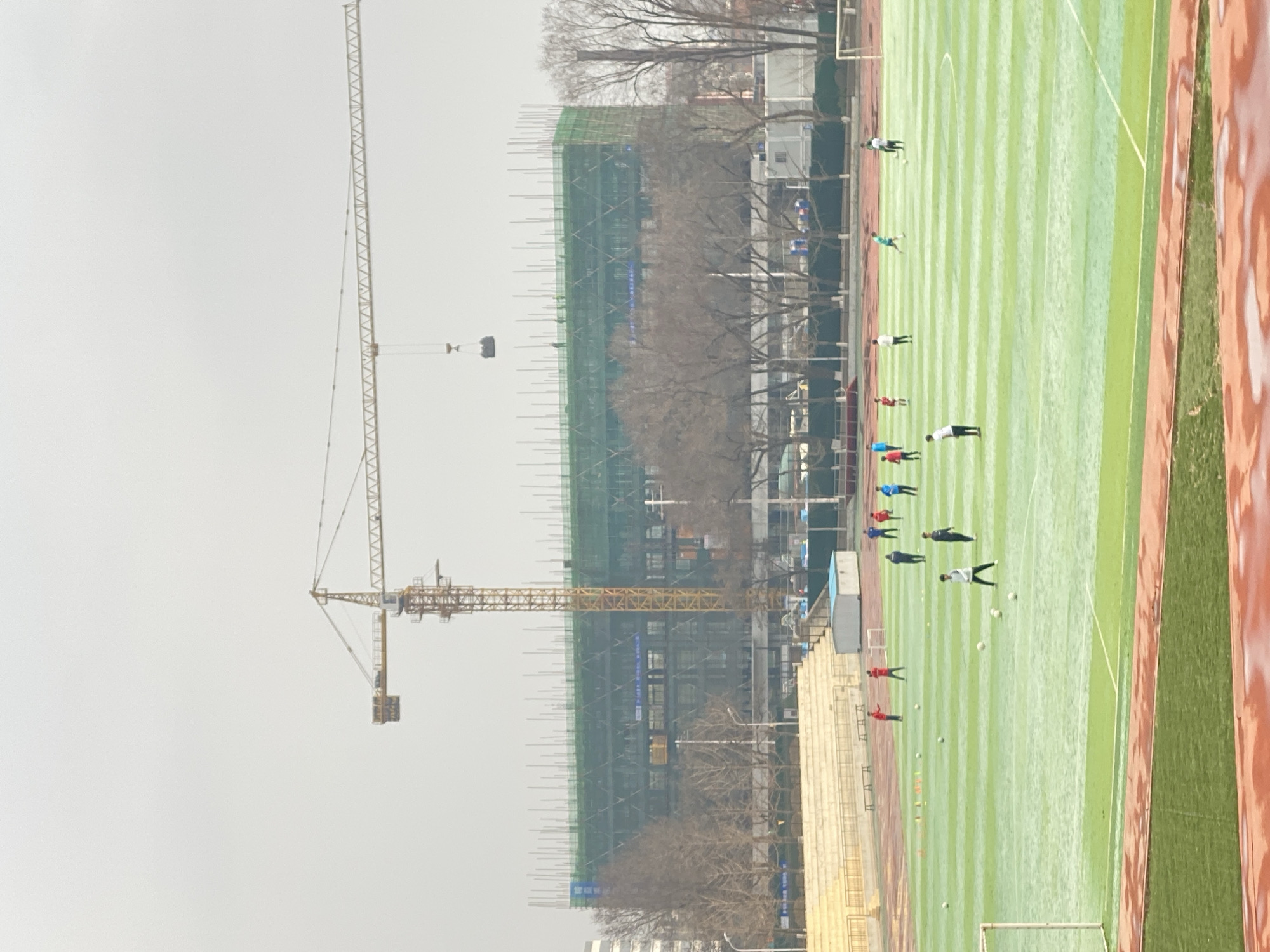 The image depicts a construction site adjacent to a sports field in Zhongtun, China. A yellow construction crane dominates the left side of the frame, its jib extending towards a building under construction, which is shrouded in green scaffolding and netting. To the right, a green artificial turf field features multiple individuals, likely athletes, engaged in a sports activity, possibly soccer or football. A running track borders the right edge of the field. The sky is overcast and grey, creating a diffused lighting effect. Trees are visible in the middle ground, adding to the urban environment.