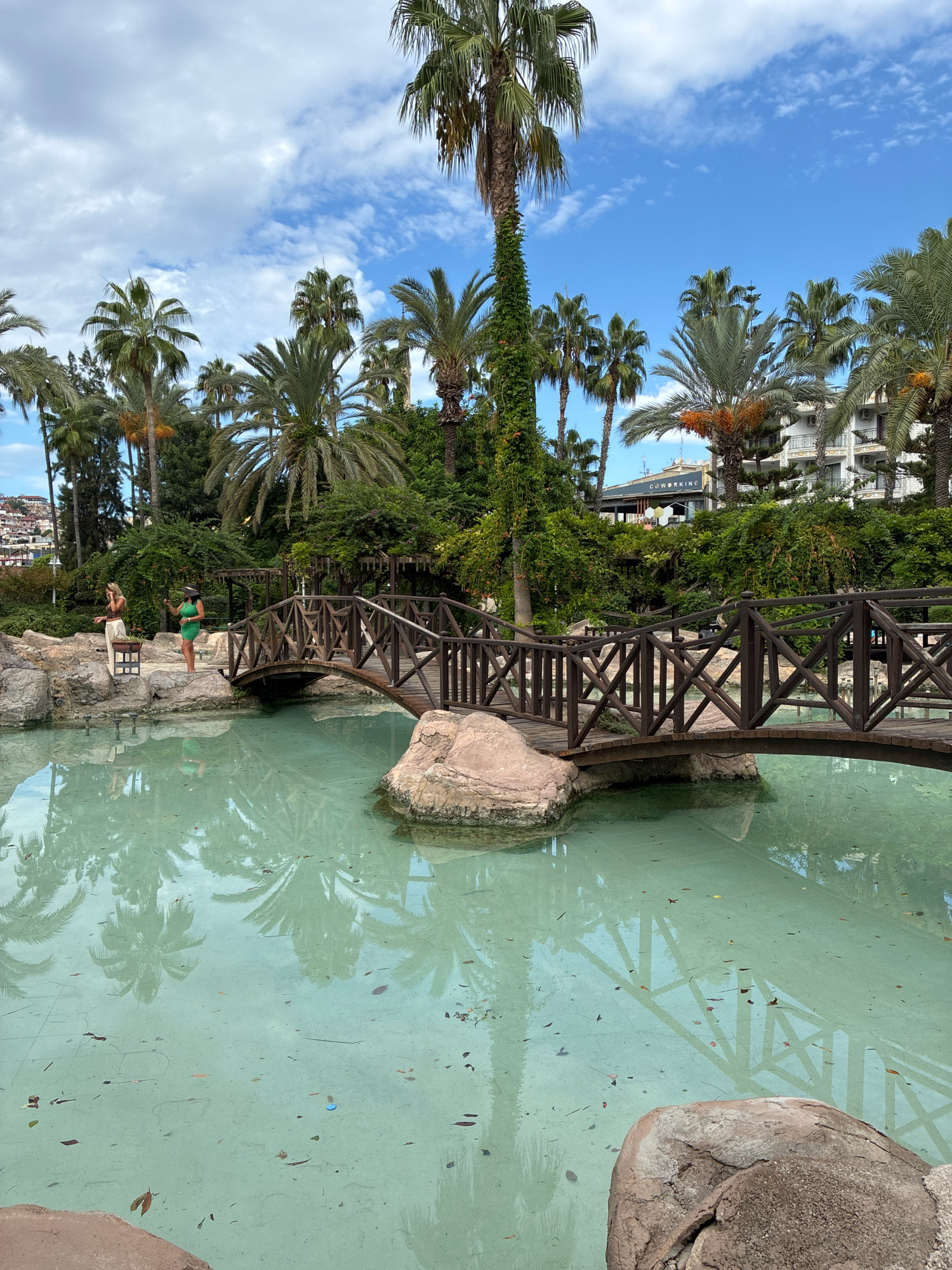 In Alanya, Turkey, a wooden bridge spans a shallow, clear turquoise pool with large rocks and palm trees surrounding it. Two women are standing on a rock ledge to the left of the bridge; one wears a green dress and the other a light-colored top and pants. Lush green foliage and palm trees line the background, with buildings visible in the distance. The sky is blue with scattered clouds.