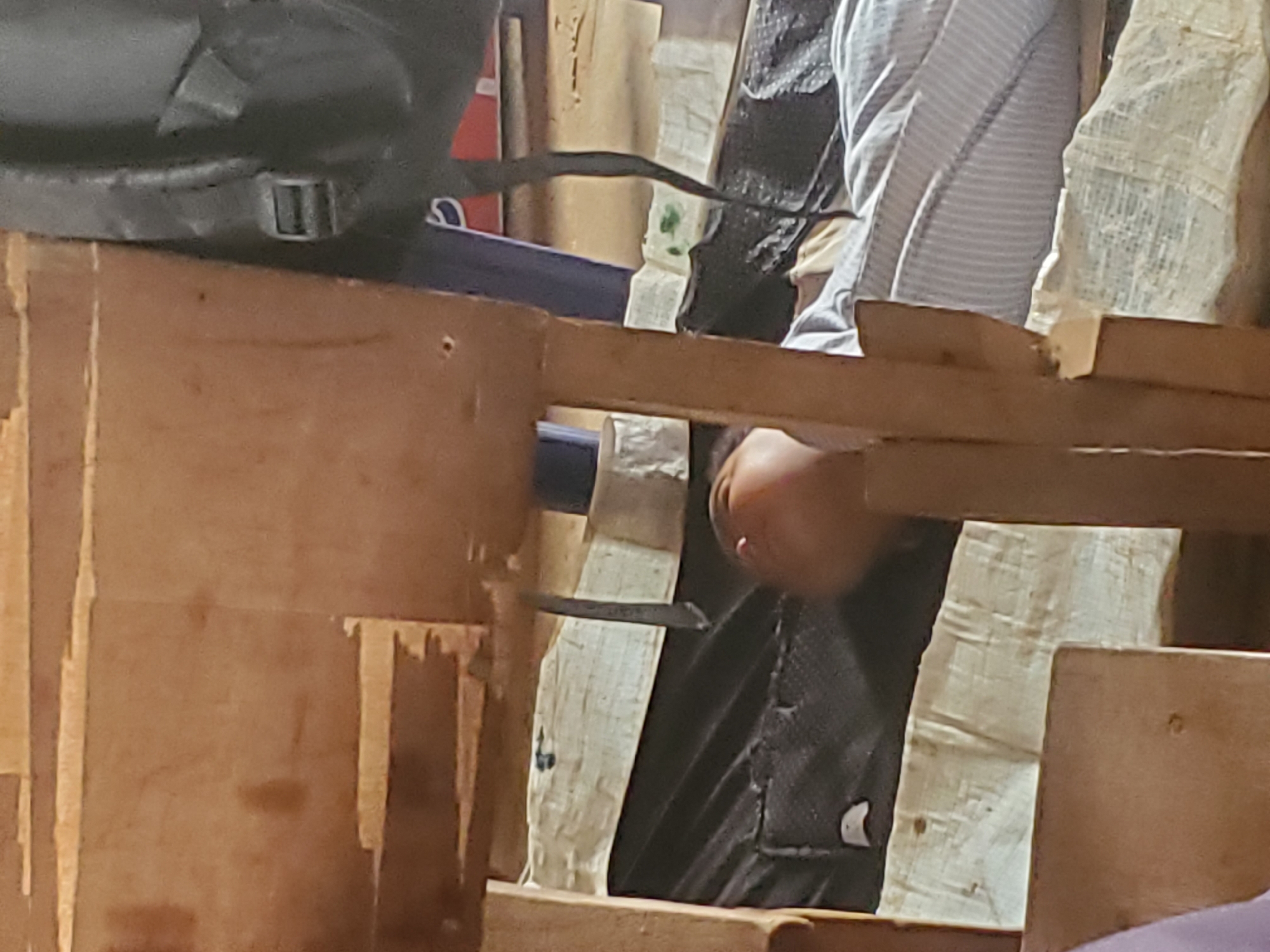The image shows a close-up of a person using a chisel to work on a wooden structure. The person is wearing a striped shirt and dark pants. A backpack is visible on the left side of the frame. The wooden structure appears to be constructed from rough-cut lumber. A hand is visible holding the chisel, which is being used to remove material from a piece of wood. The background includes some fabric and additional pieces of wood. The location is not identifiable as Ukwu-Abwa, Nigeria, from the visual information provided.