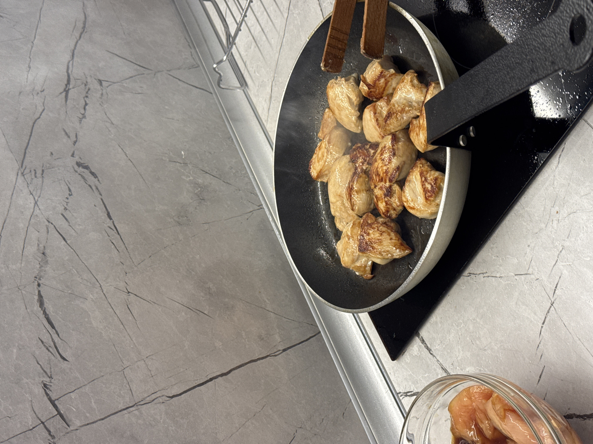 The scene depicts a top-down view of food preparation on a kitchen counter. A frying pan sits on a black stovetop, filled with pieces of cooked chicken being manipulated with wooden tongs. Steam rises from the pan, indicating recent cooking activity. To the lower right, a glass bowl contains raw chicken marinated in a dark sauce. The countertop has a light grey, veined pattern resembling marble. A dish rack is visible on the left side of the frame. No people are visible in the image. The location of the scene is unknown.