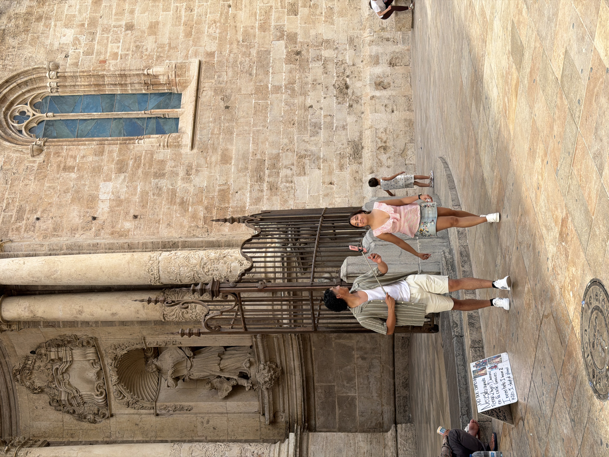 In an outdoor urban setting, a man and a woman walk past a large, ornate building with a textured stone facade and a gothic-style window. The man, on the left, wears a striped short-sleeved shirt over a white tank top and light-colored shorts, holding a selfie stick. The woman, to his right, is in a pink patterned top and a floral print skirt. Behind them, a child in a light-colored dress walks away from the camera, and further in the background, another person crouches. In the foreground, a sign with text and images is propped up on the ground, and a person sits to the left of the sign, holding a cup. The ground is paved with large, square tiles.