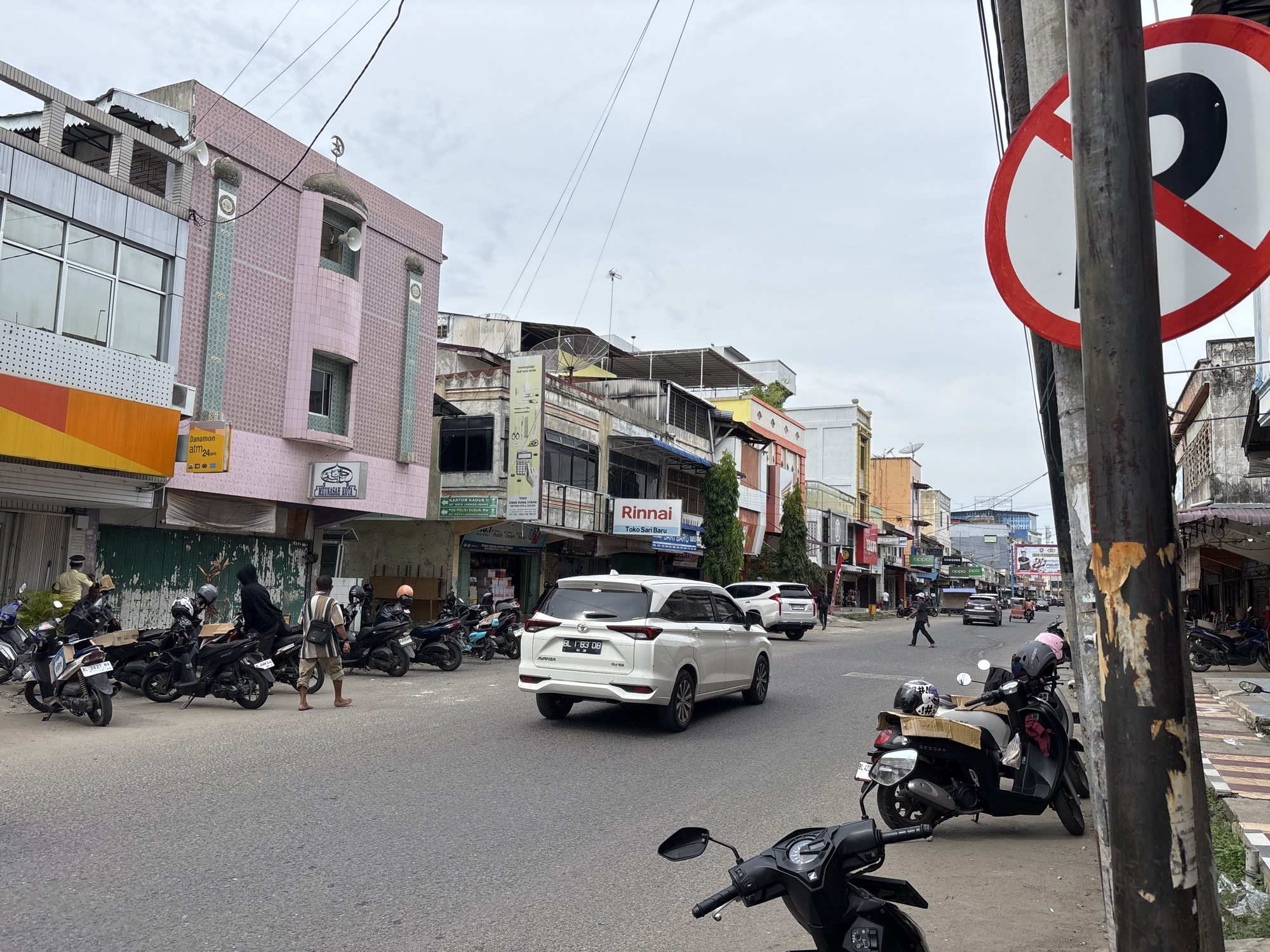 The image depicts a street scene in Lhokseumawe, Indonesia. In the foreground, several parked motorcycles are visible, some with helmets placed on them. A white SUV is driving away from the camera in the middle of the street. On the left side of the street, there are buildings with shops and signs, including a pink building with Islamic architectural elements. On the right side of the street, there are more buildings and a "no parking" sign. People are seen walking and standing on the sidewalks and in the street. The environment appears to be an urban commercial area with a cloudy sky.
