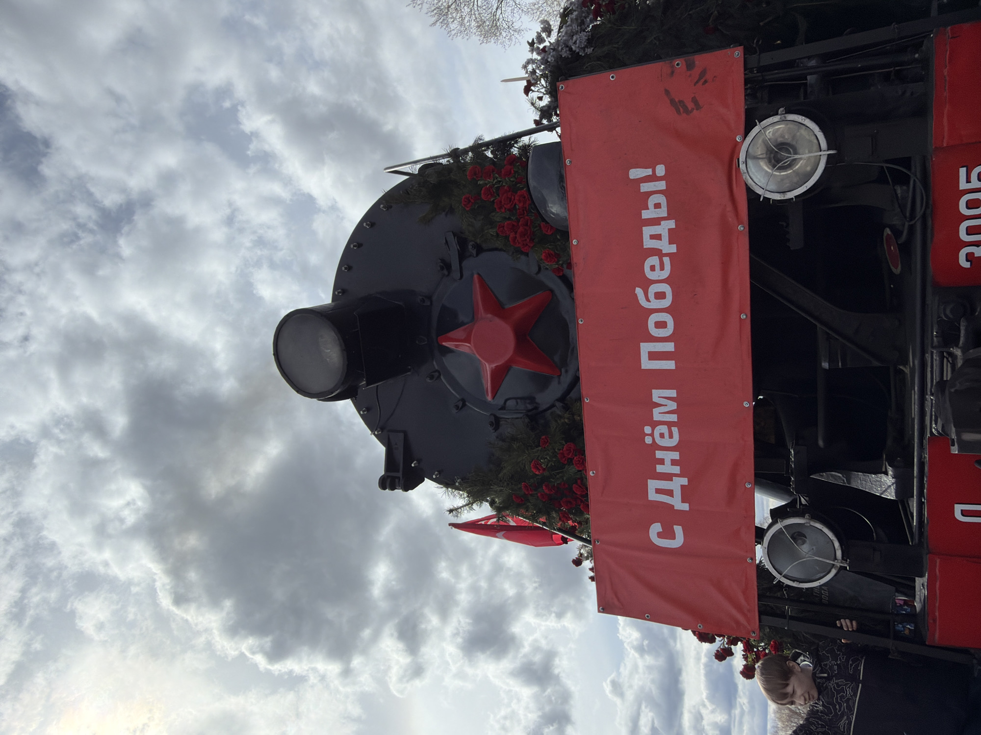 The image depicts the front of a black steam locomotive, heavily decorated for a commemorative event, likely Victory Day, in Votkinsk, Russia. The locomotive's large circular smokebox is adorned with a prominent red five-pointed star in its center and features a headlamp on top. Green foliage, resembling fir branches, and numerous artificial red roses are draped around the locomotive's front, extending above and around the main banner. A smaller red flag is also visible among the decorations.
A large red banner stretches horizontally across the front of the locomotive, bearing the Cyrillic inscription "С Днём Победы!" (S Dnyom Pobedy!), which translates to "Happy Victory Day!". To the right of the banner, a red section with the numbers "3005" is partially visible, likely part of the locomotive's identification or a connected structure.
A young person, possibly a boy, with light hair and wearing a dark patterned jacket, is visible on the lower right side of the frame, standing near the locomotive. The scene is outdoors under a cloudy, overcast sky, indicating daytime conditions. The overall impression is one of solemn celebration and remembrance.