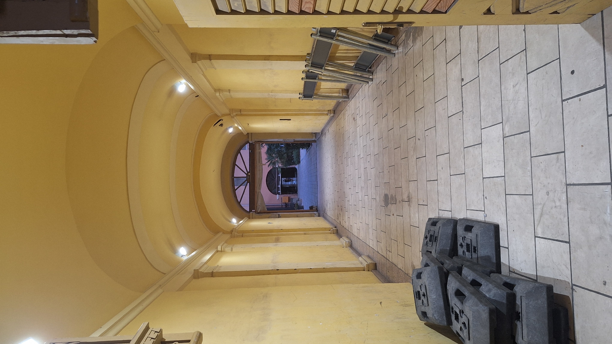 An arched, covered walkway with yellow walls and ceiling lights illuminated. The walkway is paved with light-colored rectangular tiles. On the left, near the foreground, is a stack of black, rectangular weights. On the right, towards the middle ground, are several gray metal folding barriers. At the end of the walkway, an open archway reveals a courtyard with a person standing in the distance. The exterior of the building facing the courtyard appears to be a reddish-orange color, and there are some plants visible. The scene suggests an outdoor space in a European city, possibly a public square or a private courtyard accessible from a street. The lighting indicates it is either dusk or nighttime.