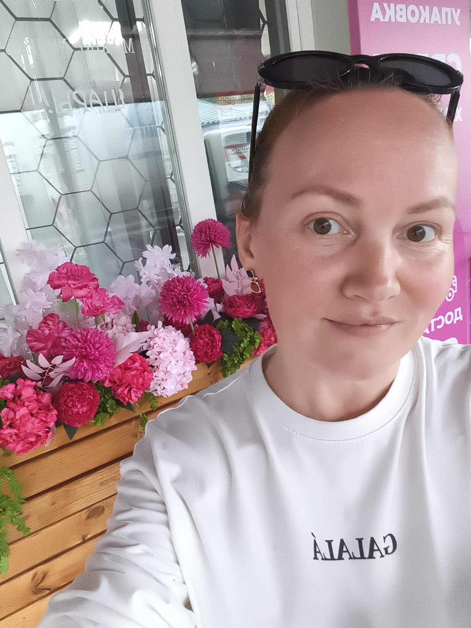 A selfie shows a woman with blonde hair and sunglasses perched on her head, looking at the camera. She is wearing a white t-shirt with a dark, stylized text graphic on it. She is framed by a window with a hexagonal pattern and a planter filled with pink and magenta flowers. In the background, pink signage with Cyrillic text is visible. The location is identified as being in the city of Chaikovsky, Russia.