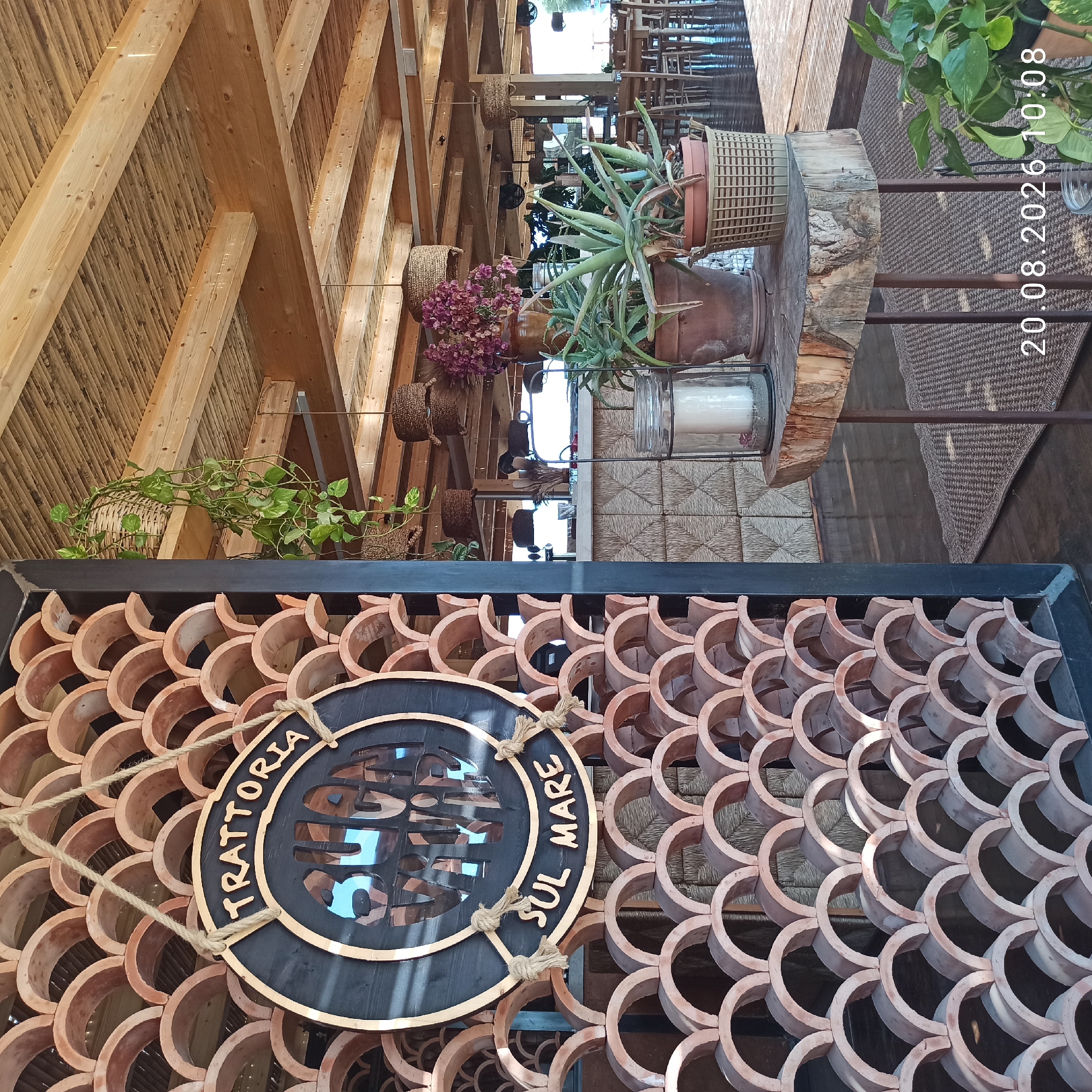 The image shows the entrance to a restaurant in Molfetta, Italy, called "Trattoria Sul Mare". A circular wooden sign with the restaurant's name hangs from a rope in front of a decorative screen made of terracotta tiles. Inside, the restaurant has a rustic, natural theme with exposed wooden beams and bamboo ceiling. Plants in woven baskets hang from the ceiling and on tables, adding to the ambiance. The date and time "20.08.2026 10:08" is superimposed on the image.