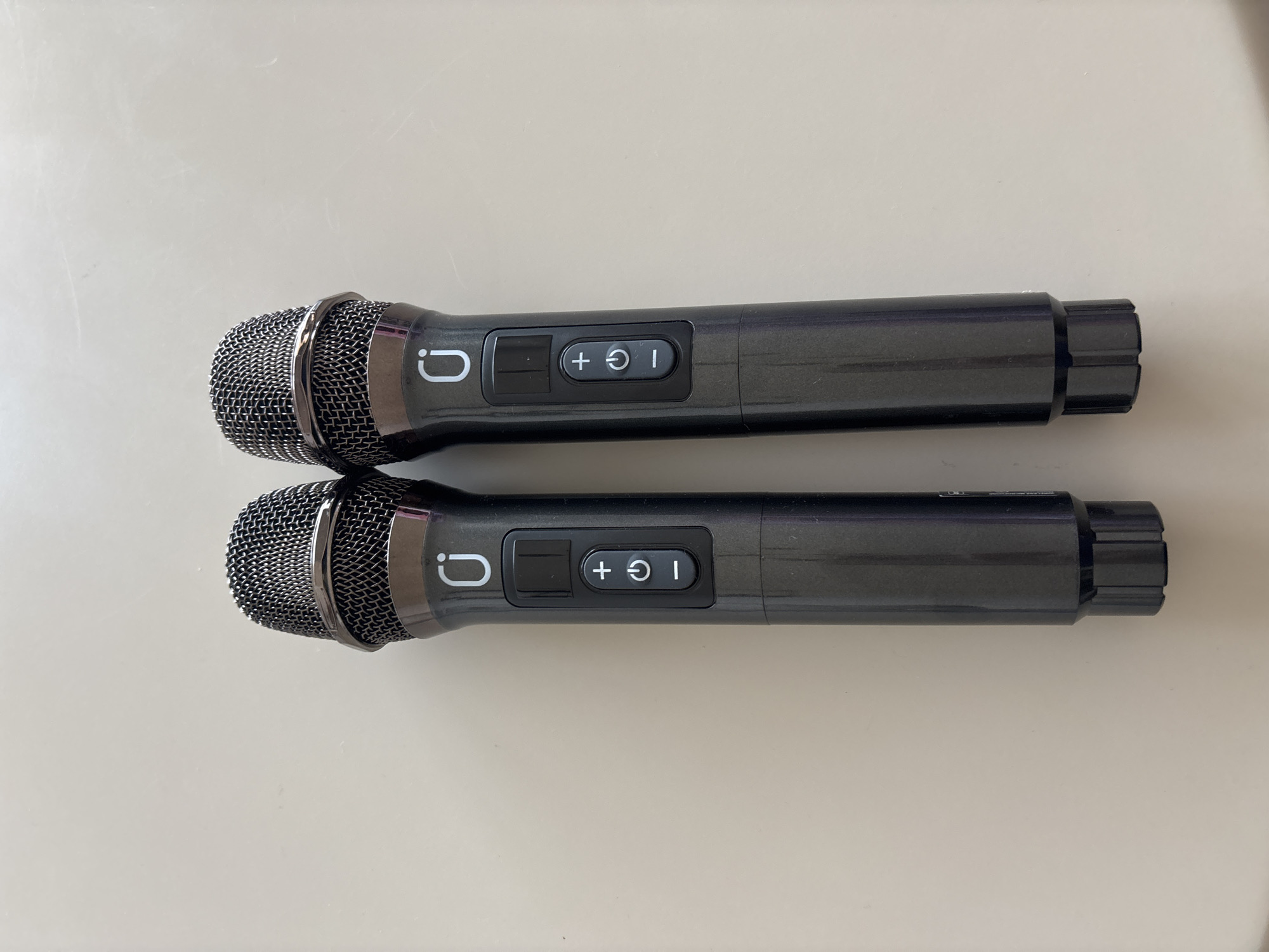 Two dark gray wireless microphones are placed side-by-side on a light-colored, flat surface, likely a table or countertop. The microphones have black mesh grilles at the top and dark gray bodies with a metallic sheen. Each microphone has a control panel with three buttons: a rectangular button at the top, a button with a plus sign and what appears to be a power symbol below it, and a button with a minus sign at the bottom. The logo of a white circular icon with a line through it is present on the body of each microphone, above the control panel. The lighting suggests an indoor environment, possibly in Dubai, United Arab Emirates, though no specific landmarks or distinguishing features of Dubai are visible. The image is a top-down view, focusing on the two microphones.