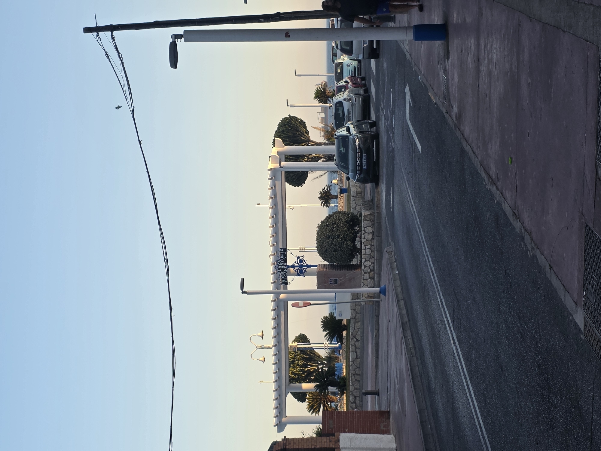 The image depicts a street scene in Málaga, Spain, likely during the late afternoon or early evening. The sky is clear and a pale blue, fading to a lighter hue near the horizon. A streetlamp with a curved arm and two lights is visible on the left side of the frame. Power lines stretch across the upper portion of the image, supported by a wooden pole on the right.
In the middle ground, there is a white pergola structure with supporting columns. Beneath the pergola, plants and trees are arranged in garden beds, with stone walls bordering the landscaped area. A street sign with blue lettering on a white background reads "PLAZA PADRE ANTONIO SEGURA." A red and white circular traffic sign is mounted on a pole near the pergola.
A row of cars is parked or driving on the street. These include a dark SUV in the foreground, followed by several smaller cars and vans. The street has white lane markings and a directional arrow painted on the asphalt. The sidewalk on the right side of the street is paved and has a reddish-purple hue. A lamppost with a white pole and blue base stands on the sidewalk.
A person is walking on the sidewalk on the far right, wearing dark shorts and a dark shirt. Another person is visible in the distance, sitting near the beachfront. The sea is visible in the background, with a ship on the horizon. The overall impression is a coastal urban setting with elements of public space, transportation, and natural elements.