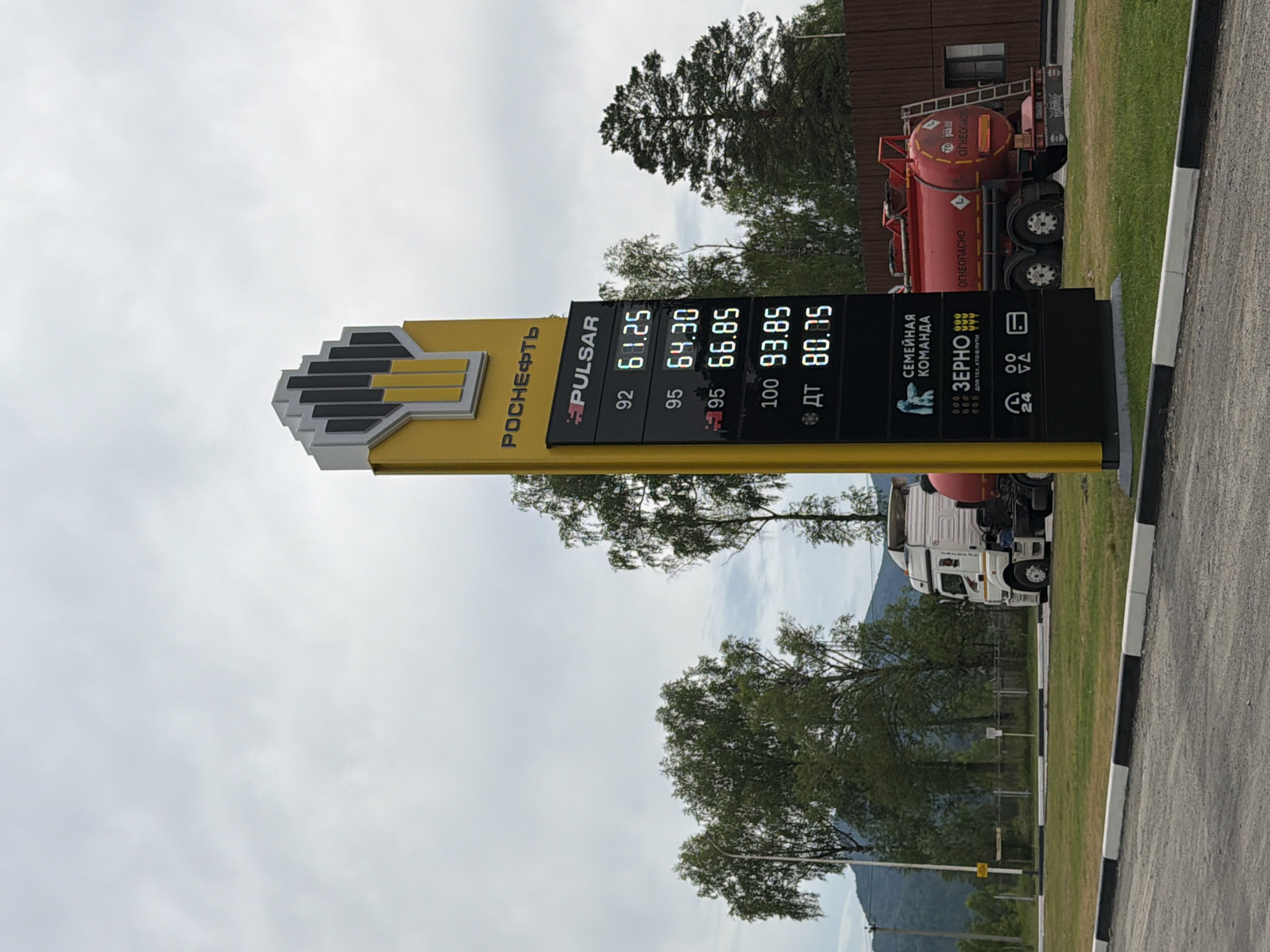 The image shows a "Rosneft" gas station in Ust'-Sema, Russia. A yellow sign displays fuel prices, with 92 octane at 61.25 rubles, 95 octane at 64.30 rubles, 95+ at 66.85 rubles, 100 octane at 93.85 rubles, and diesel (ДТ) at 80.85 rubles. Below the fuel prices, there are logos for "Pulsar," "Семейная команда" (Family Team), "Зерно" (Grain), and icons indicating 24-hour service and payment options. A white semi-truck with a red fuel tanker is parked to the right of the sign. The truck has "ОГНЕОПАСНО" (Flammable) markings. The environment is outdoor, with green grass, trees, and a cloudy sky. A road with black and white curbs is visible in the foreground.