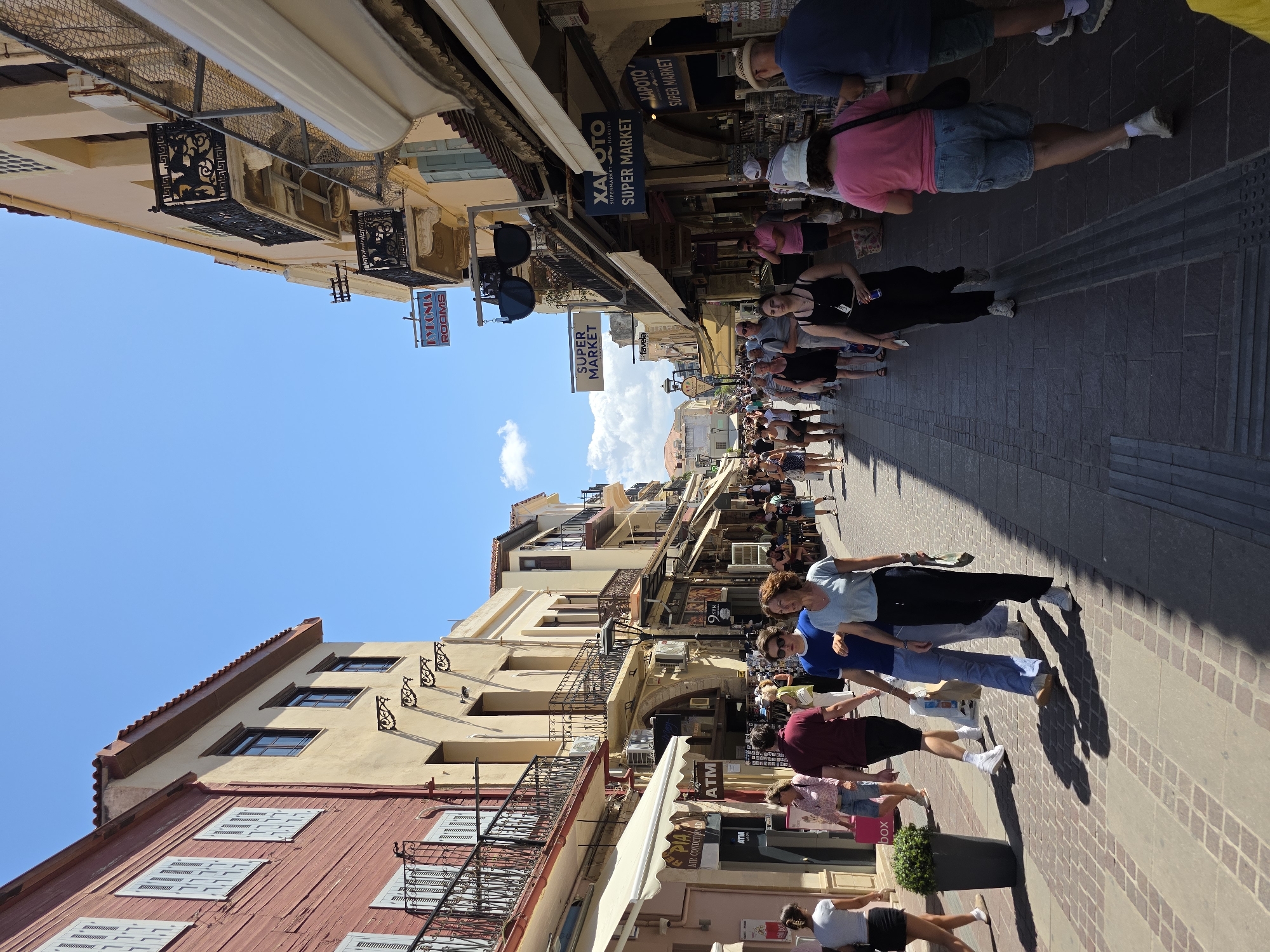 The image captures a bustling pedestrian street in Chania, Greece, likely the Old Town. The scene is dominated by narrow buildings lining both sides of the cobblestone street, their facades painted in warm, earthy tones of yellow and red, with traditional architectural details such as balconies with wrought iron railings. Shops with awnings are visible on the ground floors, with signs in Greek and English indicating businesses like "Super Market" and "ATM."
A diverse crowd of people are walking in both directions, many appearing to be tourists. They are dressed in casual summer clothing, with some carrying shopping bags. The atmosphere is lively, suggesting a popular area for shopping and sightseeing. The clear blue sky overhead, with a few scattered clouds, indicates a sunny day. The perspective of the image is from street level, looking down the length of the street, with the buildings receding into the distance. Shadows are cast across the cobblestones, indicating the sun is high in the sky.