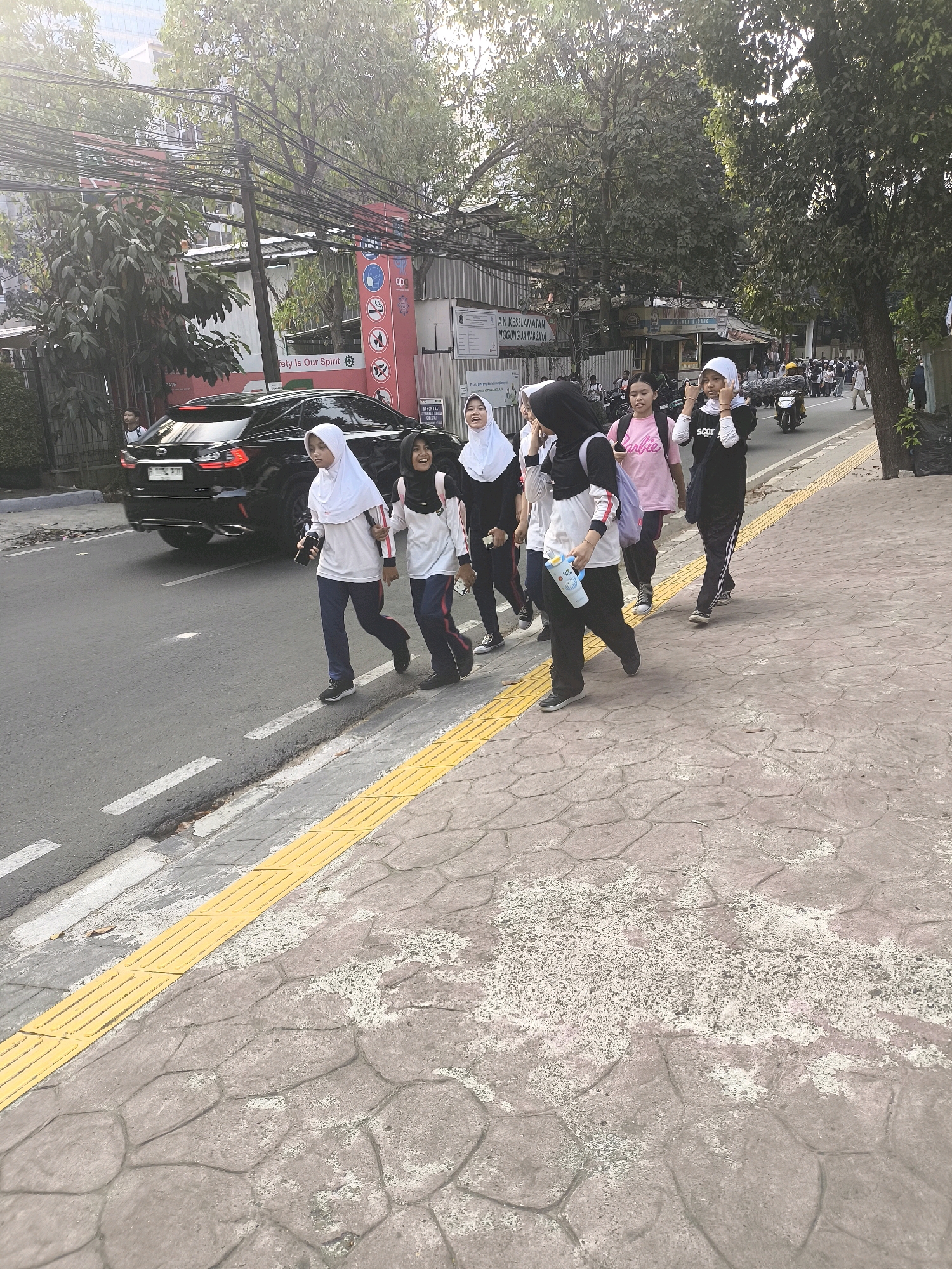 A group of six young women, all wearing headscarves, are walking on a sidewalk next to a street in South Jakarta, Indonesia. The women are dressed in casual clothing, with some wearing t-shirts and others wearing long-sleeved shirts and pants. They appear to be students, possibly on their way to or from school. One woman in the foreground is carrying a blue water bottle, and another is holding a phone. The sidewalk is paved with patterned concrete and has a yellow tactile paving strip. The street is asphalt and has white lane markings. A black SUV is driving on the street, and a few motorcycles are visible in the background. Buildings and trees line the street, suggesting an urban environment. The lighting indicates it is daytime.