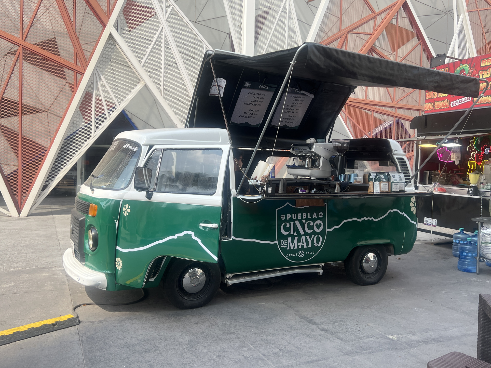 The image shows a green and white Volkswagen van converted into a mobile coffee stand, parked outdoors. The van has the words "PUEBLA CINCO DE MAYO DESDE 1862" painted on its side. Menus are displayed under the black canopy, listing drinks such as cappuccino, latte, mocha, and chai. An espresso machine and other coffee-making equipment are visible. In the background, there is a modern architectural structure with red and white triangular patterns. Another food stand is seen to the right of the van, with a sign that mentions "Mango Habanero 100% Calidad." Water bottles are stacked nearby. The image appears to be taken in a public space or plaza.